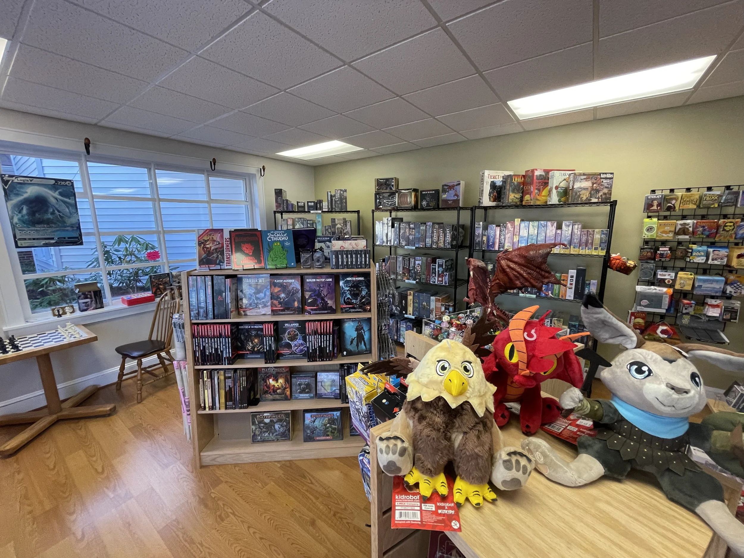 A cozy board game and toy store with shelves filled with board games, boxed miniatures, and accessories. There are plush toys on a table, including a large bird, a red dragon, and a rabbit, along with a window with blinds letting in natural light. A 