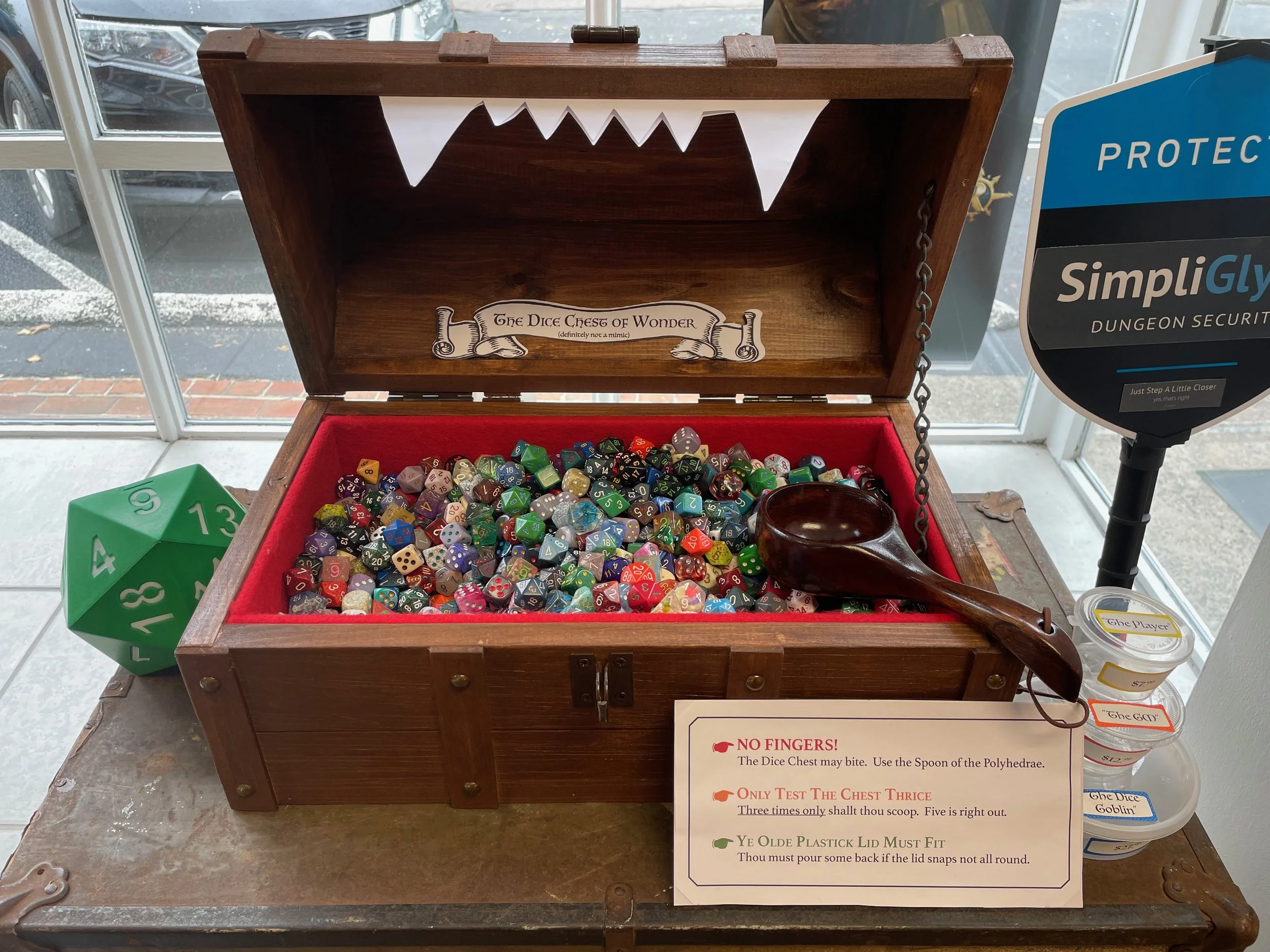 A wooden dice chest filled with an assortment of polyhedral dice used for tabletop gaming. The chest is open, showing a tufted red lining, with a sign inside reading 'The Dice Chest of Wonder.' Next to the chest is a large green 20-sided die, and on 