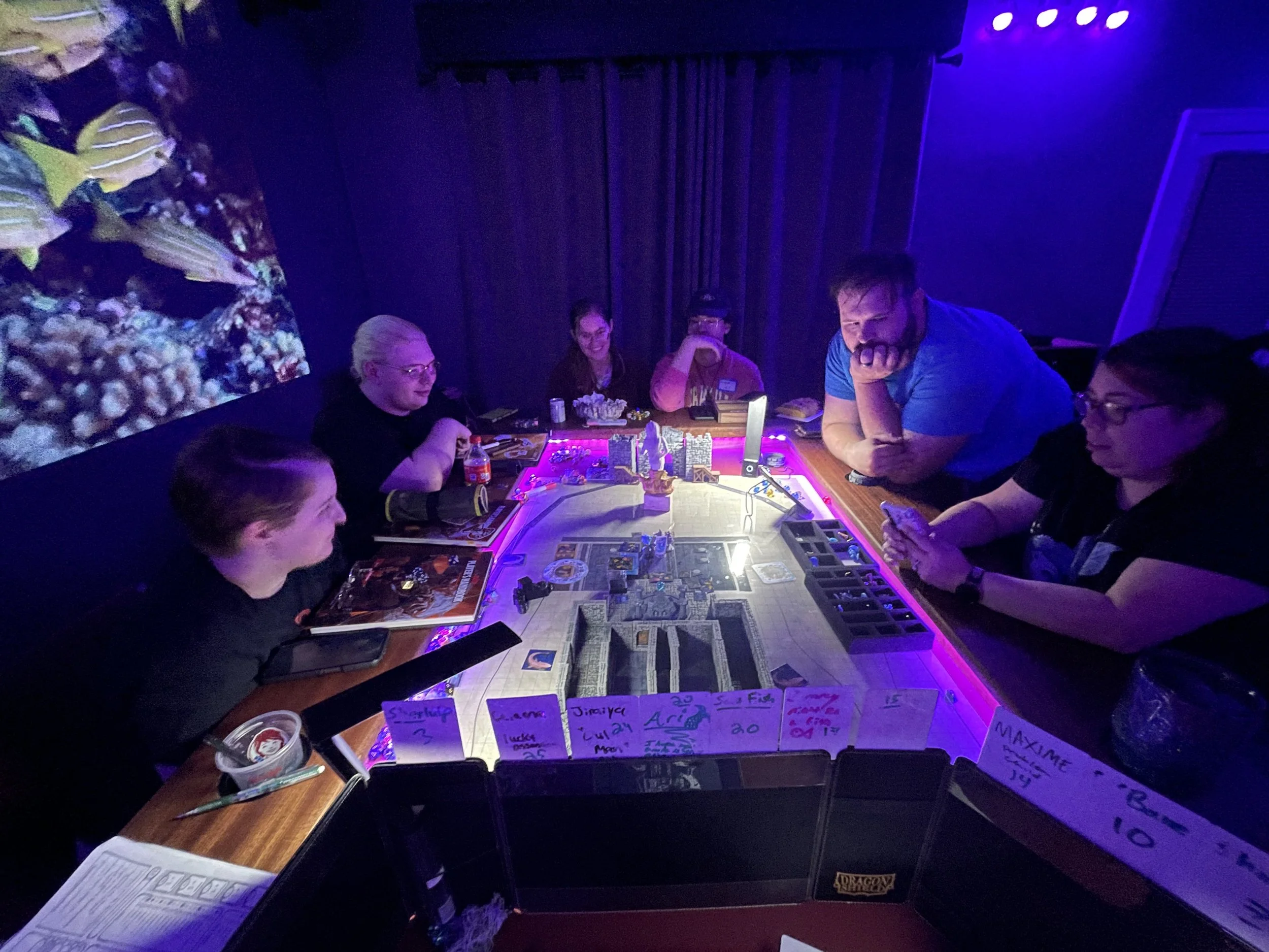 Group of six people playing a tabletop role-playing game at a table with a large game board, miniatures, and notes, in a dimly lit room with a screen displaying underwater coral scene.