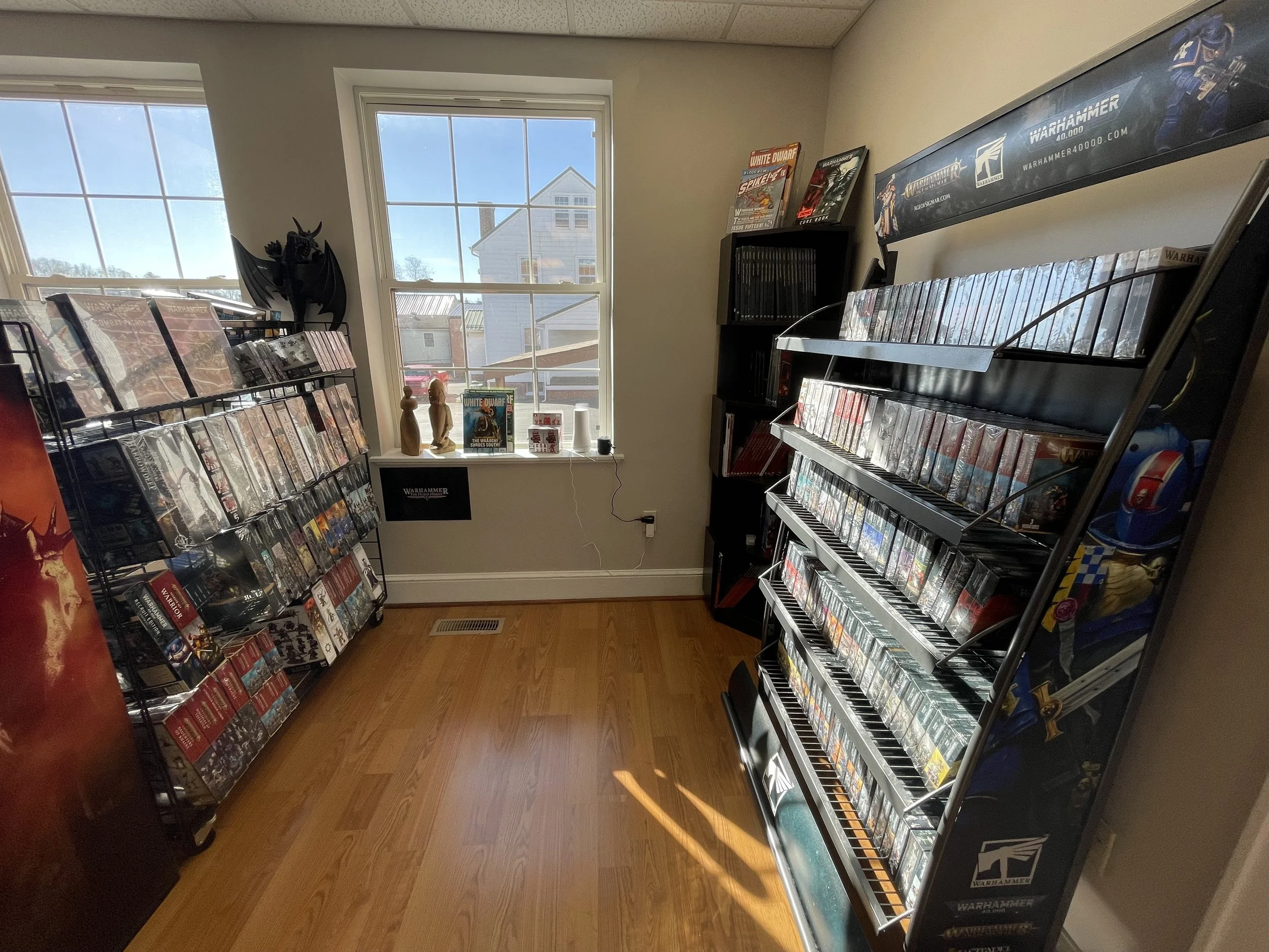 A small room with wooden flooring, two large windows, and shelves filled with Warhammer miniature games and books. There is a black dragon figurine on the left shelf, decorative wooden mannequins on the windowsill, and a poster on the wall.