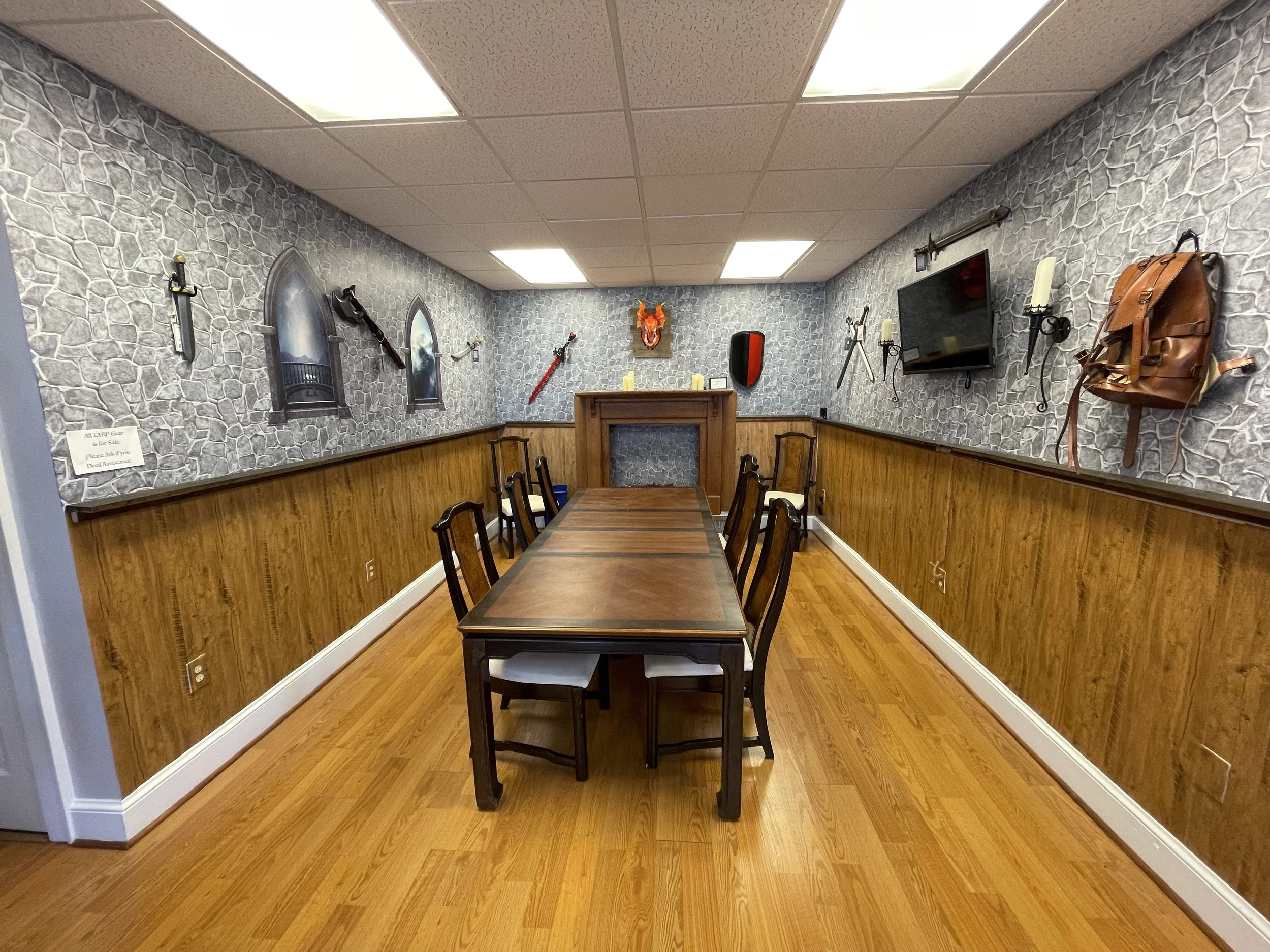 A medieval-themed dining room with a long wooden table and chairs. The wall has castle window decals, a dragon mask, swords, and a TV. The lower wall has wood paneling, and the floor is hardwood.