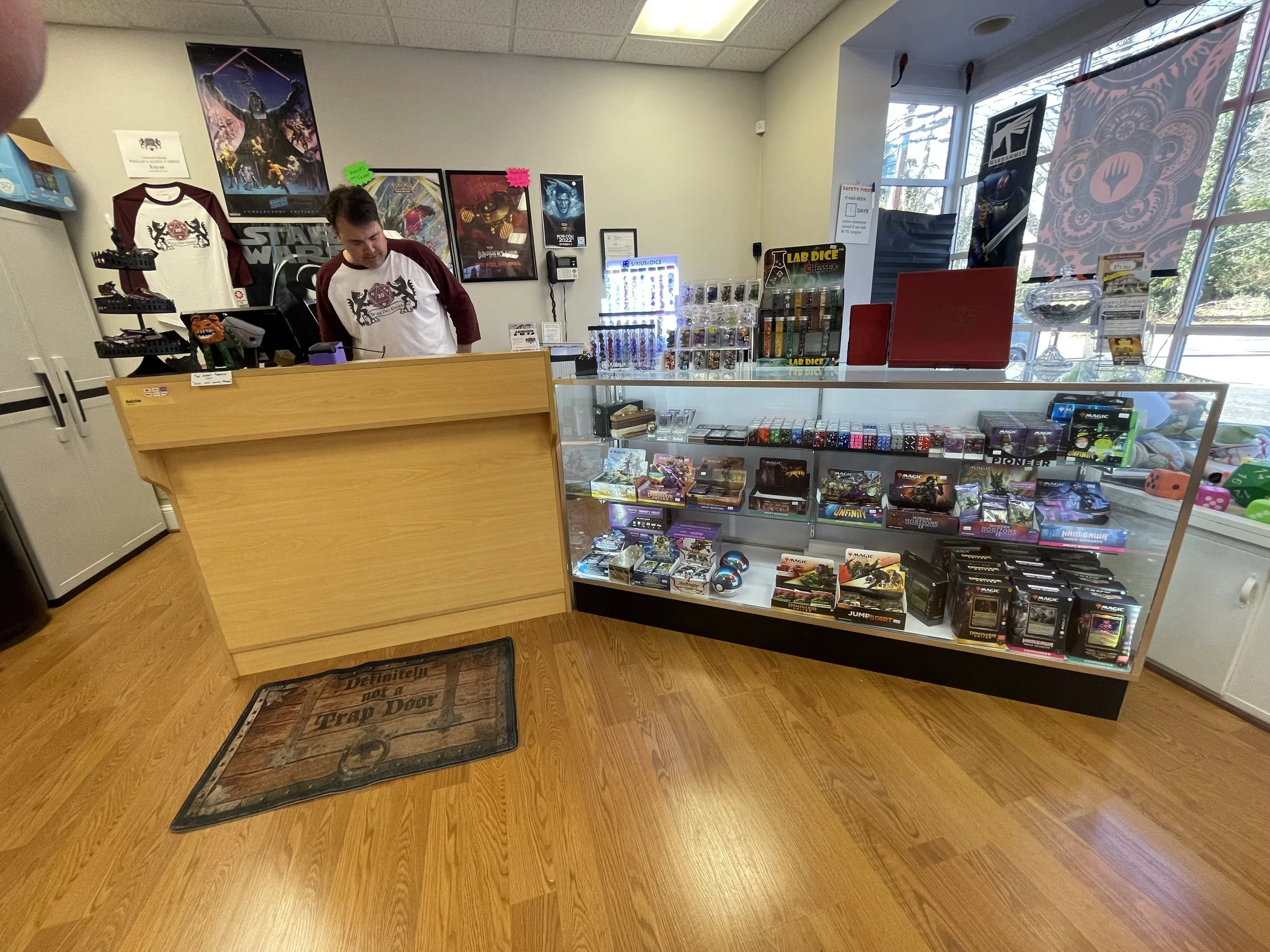 Inside a game store with a wooden counter, store employee behind it, and shelves displaying board games, card games, dice, and collectibles. Posters on the wall and a large window allowing natural light.