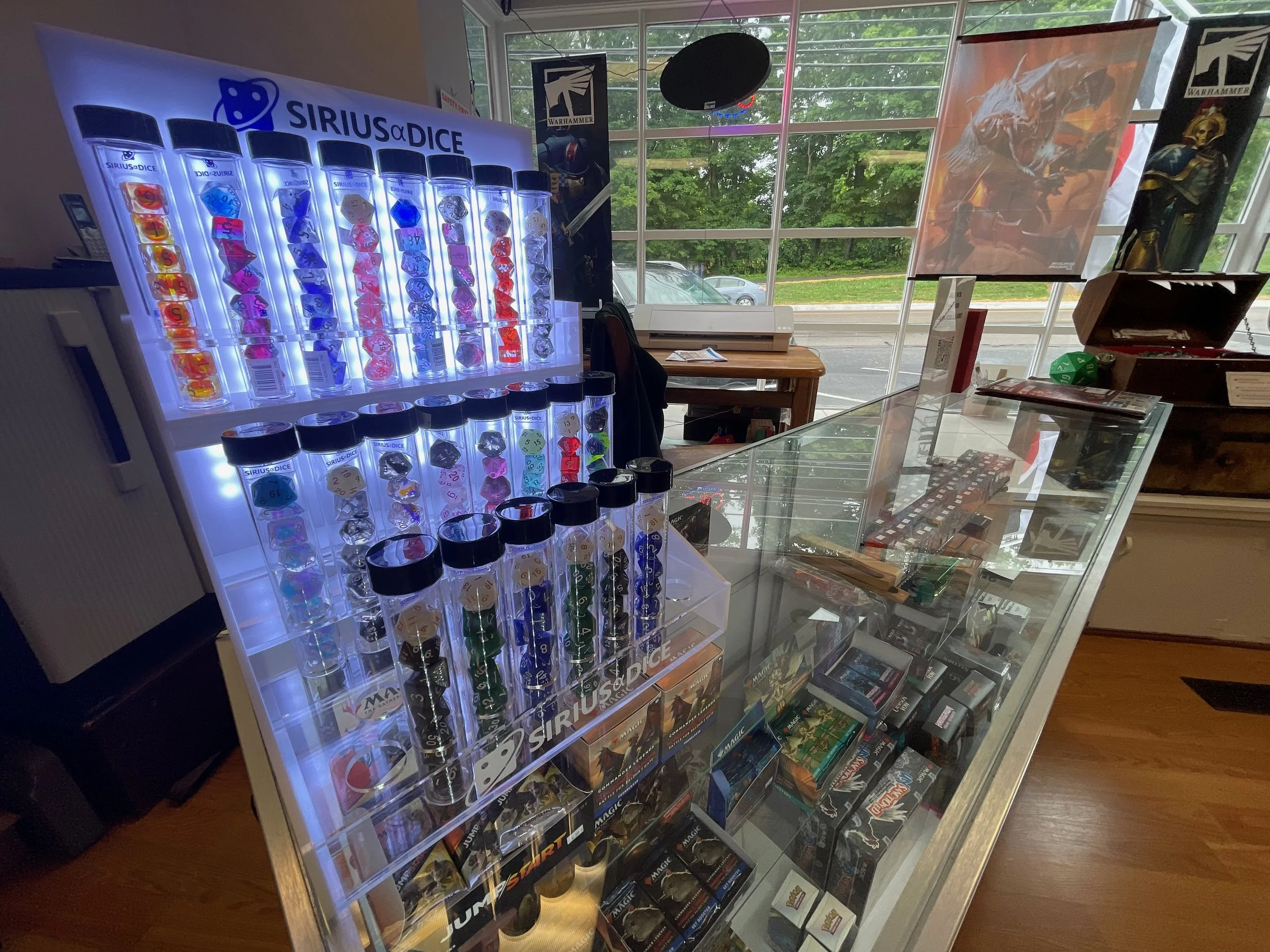 Display case with tabletop miniatures, board games, and game accessories inside a store, with a large illuminated shelf of colorful dice and game boxes, and a window showing trees and parked cars outside.