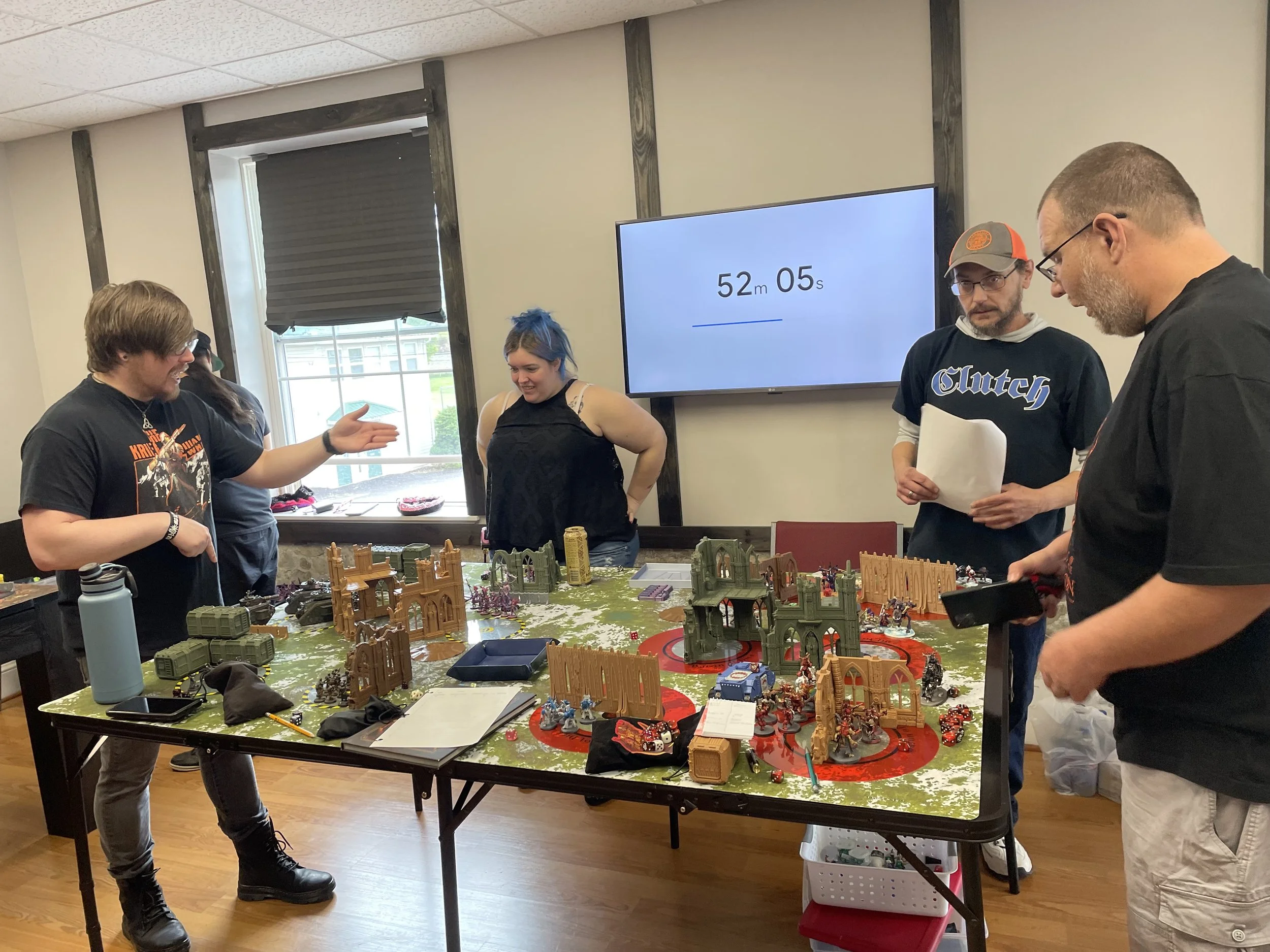 Group of five people playing a tabletop miniature war game with detailed terrain and miniatures on a large table, indoors near a window.
