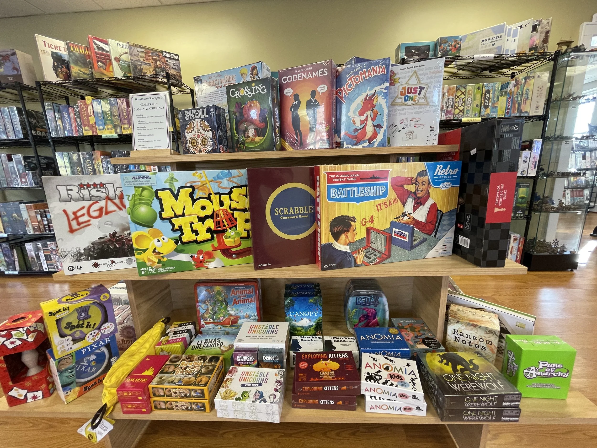 A wooden display shelf filled with various board games and puzzles, including Risk, Mouse Trap, Scrabble, Battleship, and Animal Upon Animal, in a store setting with shelves of other games and collectibles in the background.