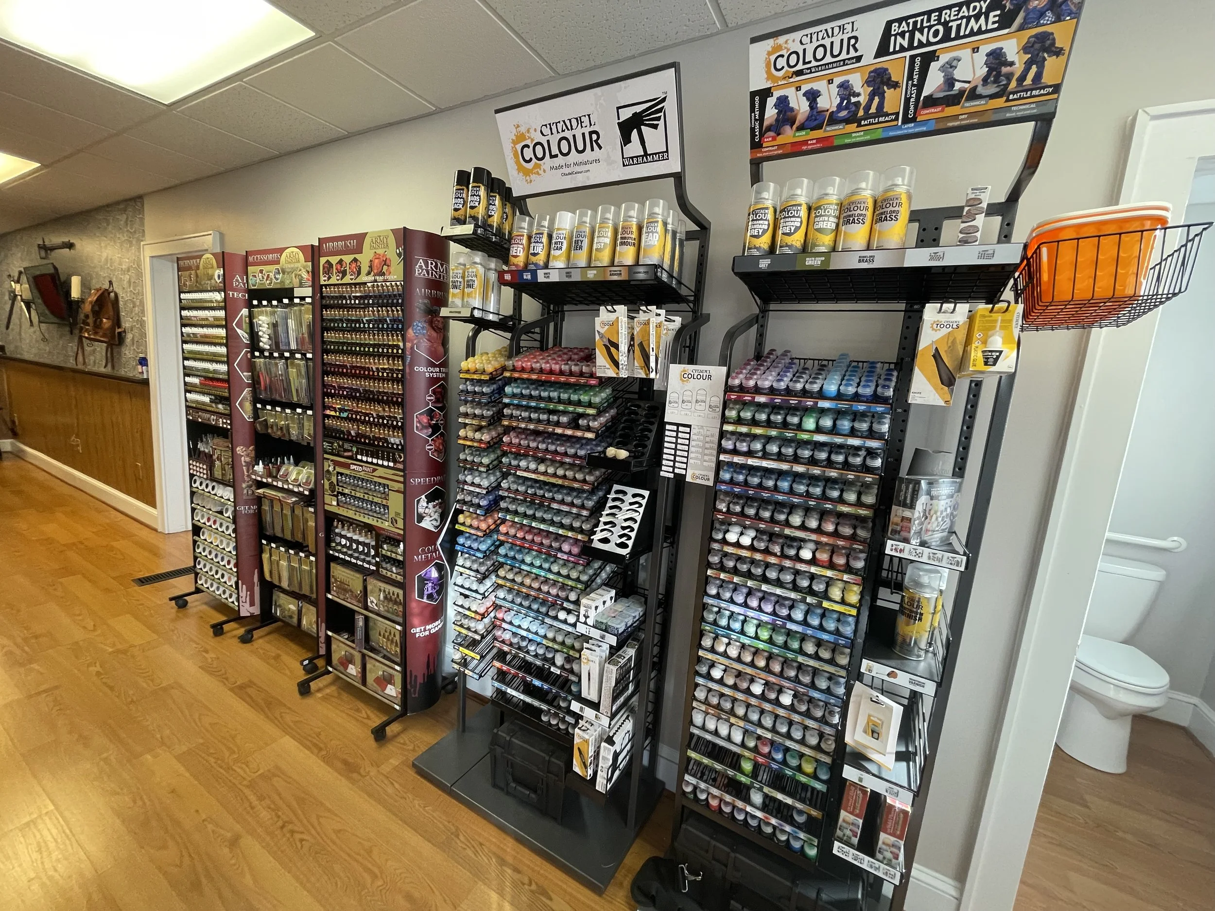 Display of Citadel miniature paints organized on black metal racks in a hobby or craft store, with a toilet partially visible in the background.