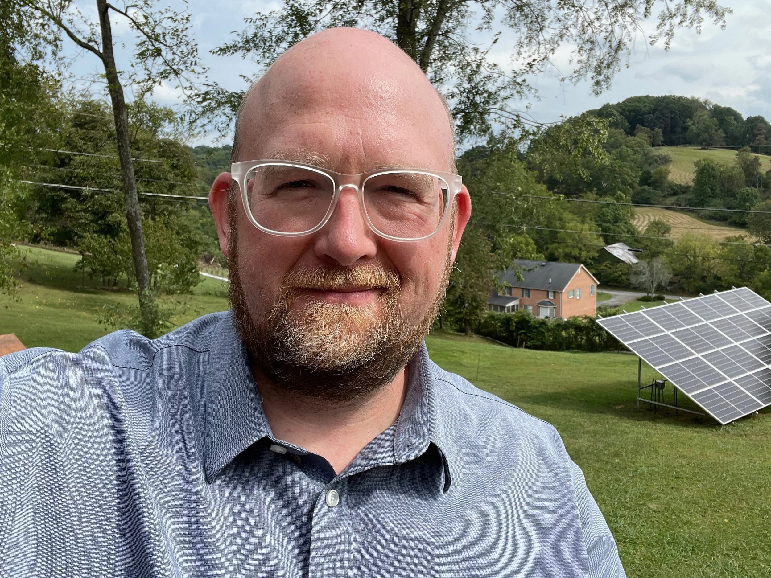 A man with glasses and a beard taking a selfie outdoors on a cloudy day, with a rural landscape including a solar panel, trees, houses, and rolling hills in the background.
