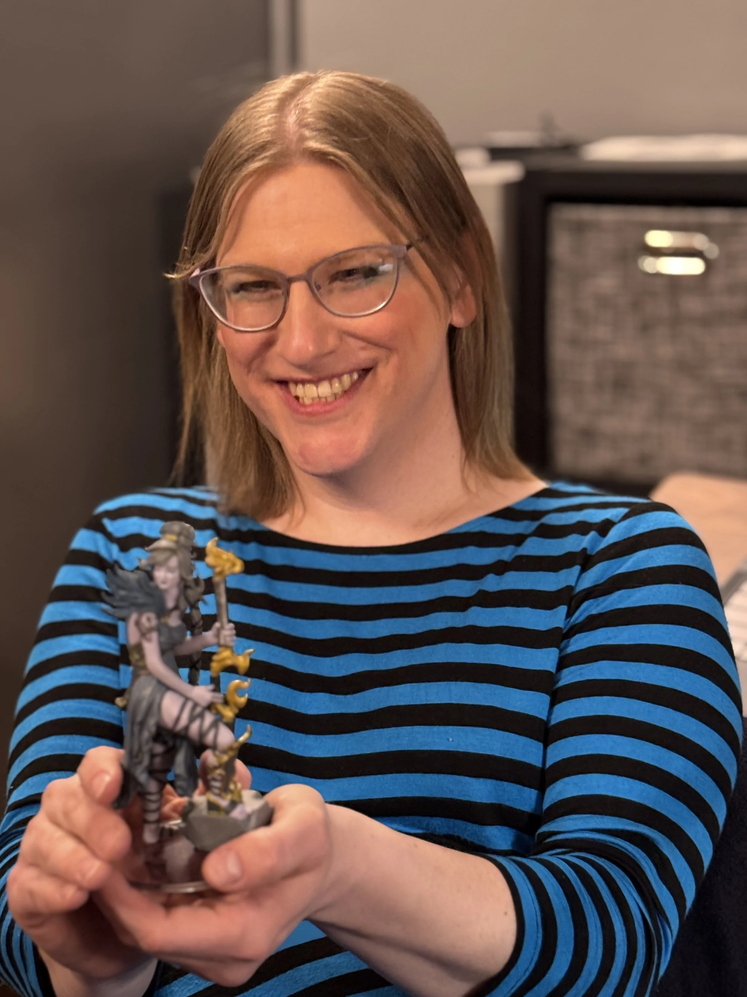 Woman with glasses wearing a blue and black striped shirt holding a fantasy miniature figurine.