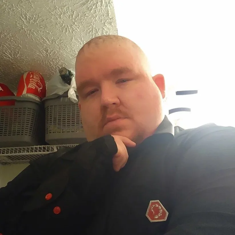 A portrait of a man with short hair wearing a black shirt with insignia.