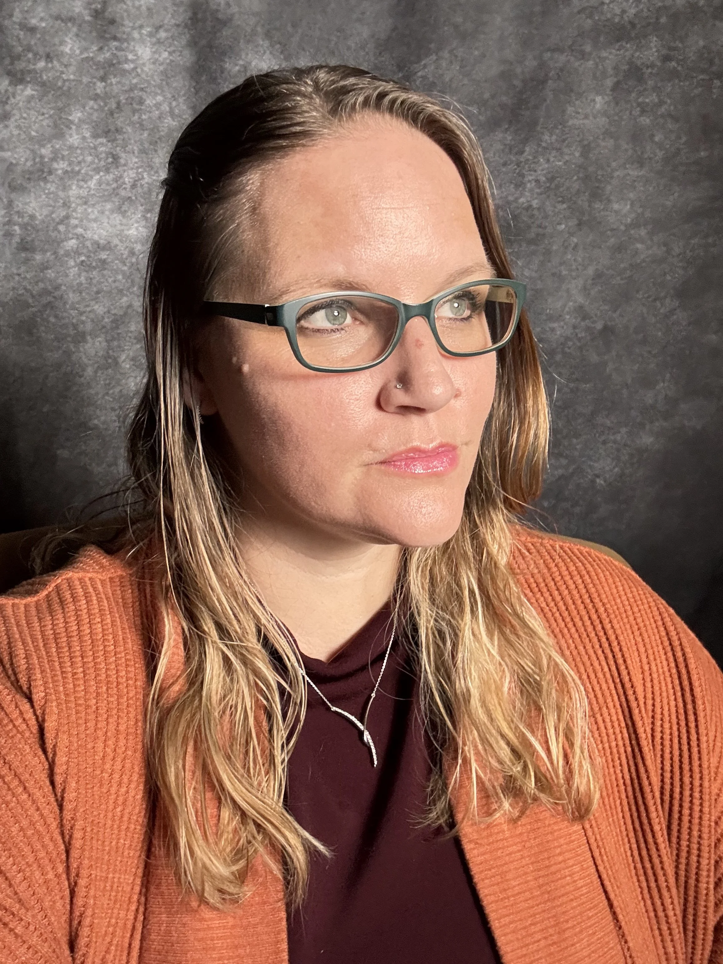 Close-up of a woman with long, wavy hair, wearing glasses, a nose piercing, a necklace, an orange sweater, and a maroon top, against a dark textured background.