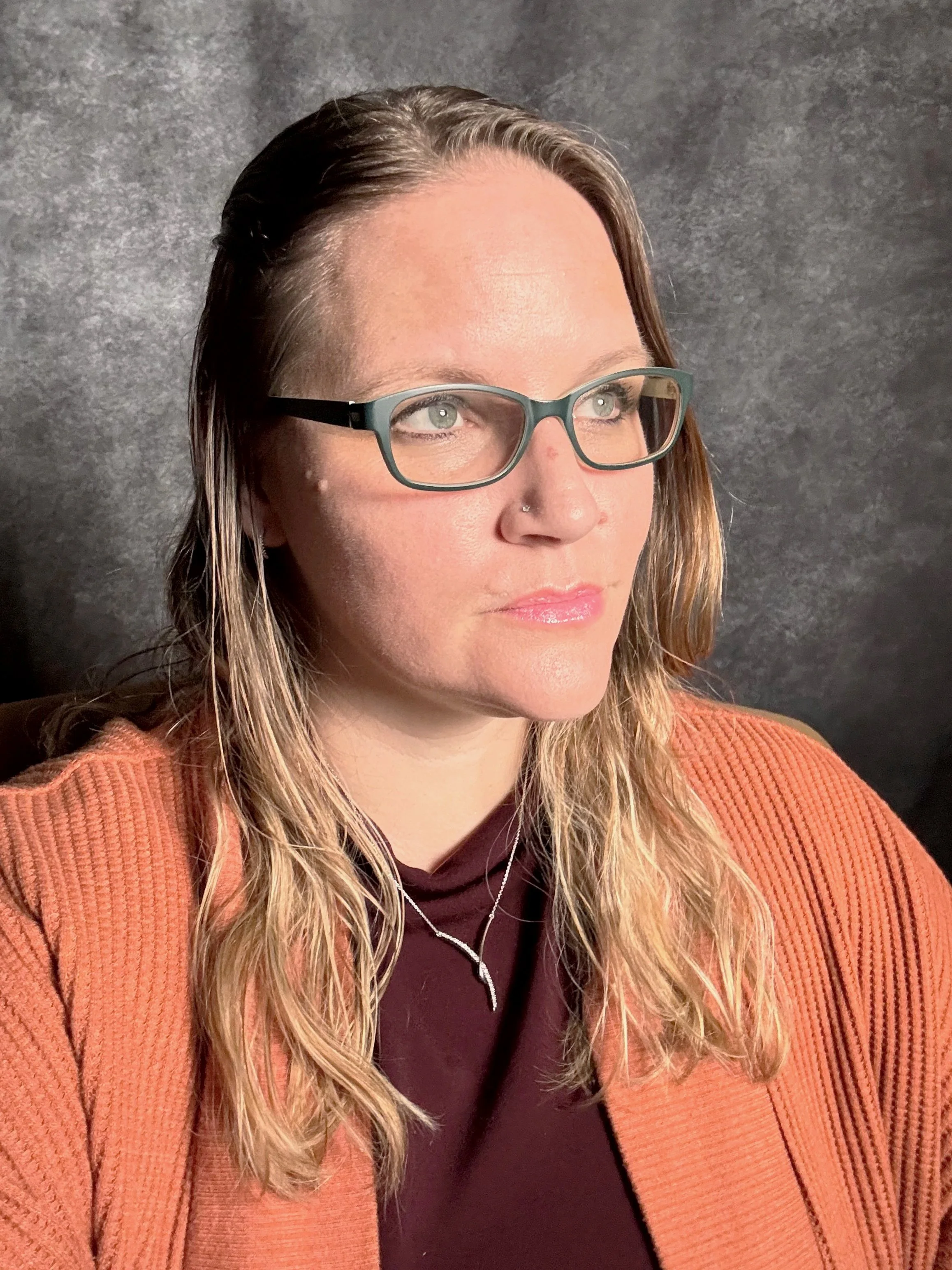 A woman with long wavy hair wearing glasses, an orange ribbed sweater, a black shirt, and a layered necklace, looking slightly to the right with a neutral expression.