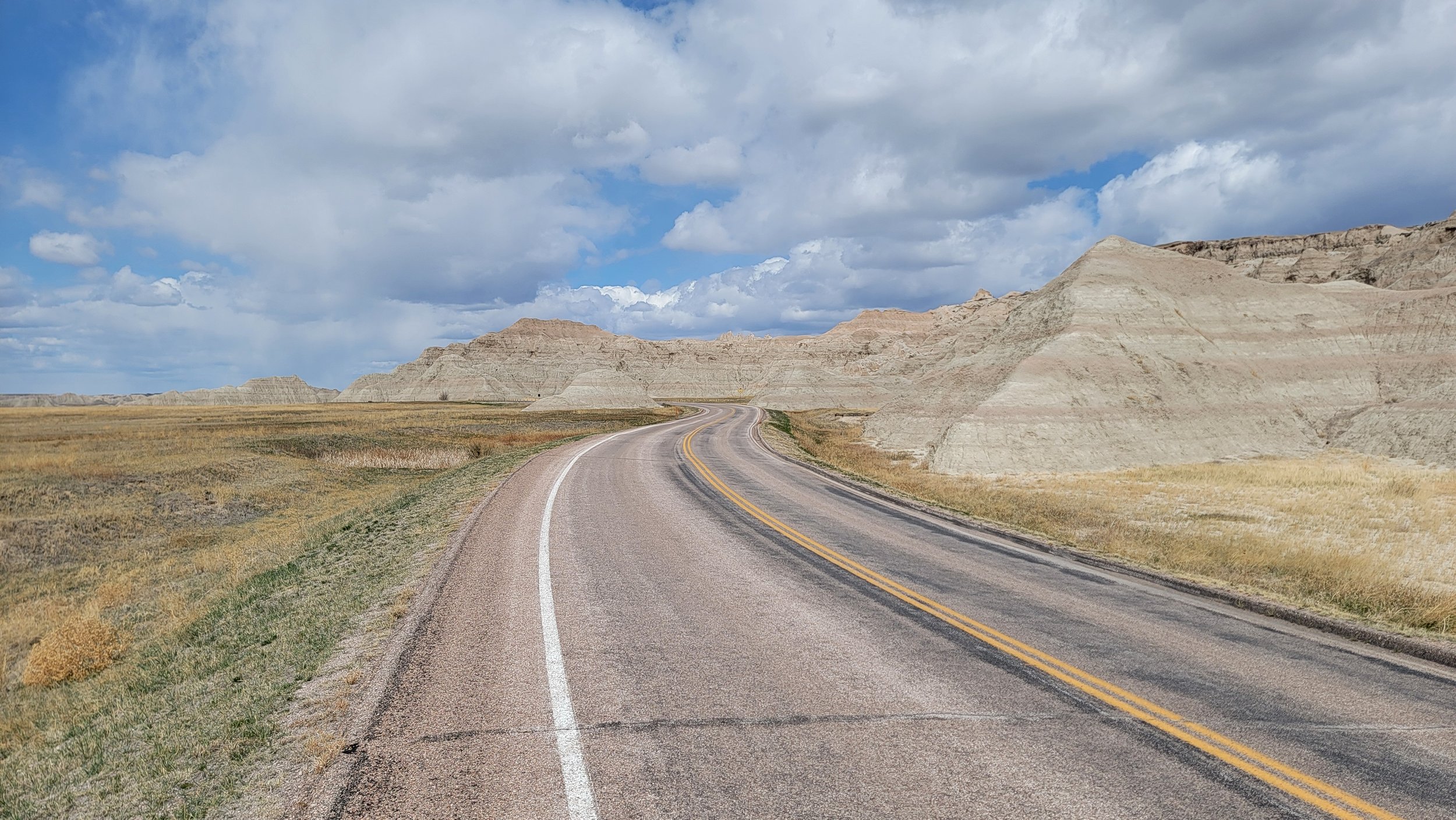 A winding two-lane road cutting through a flat, open landscape with layered, light-colored rock formations and a partly cloudy sky overhead.