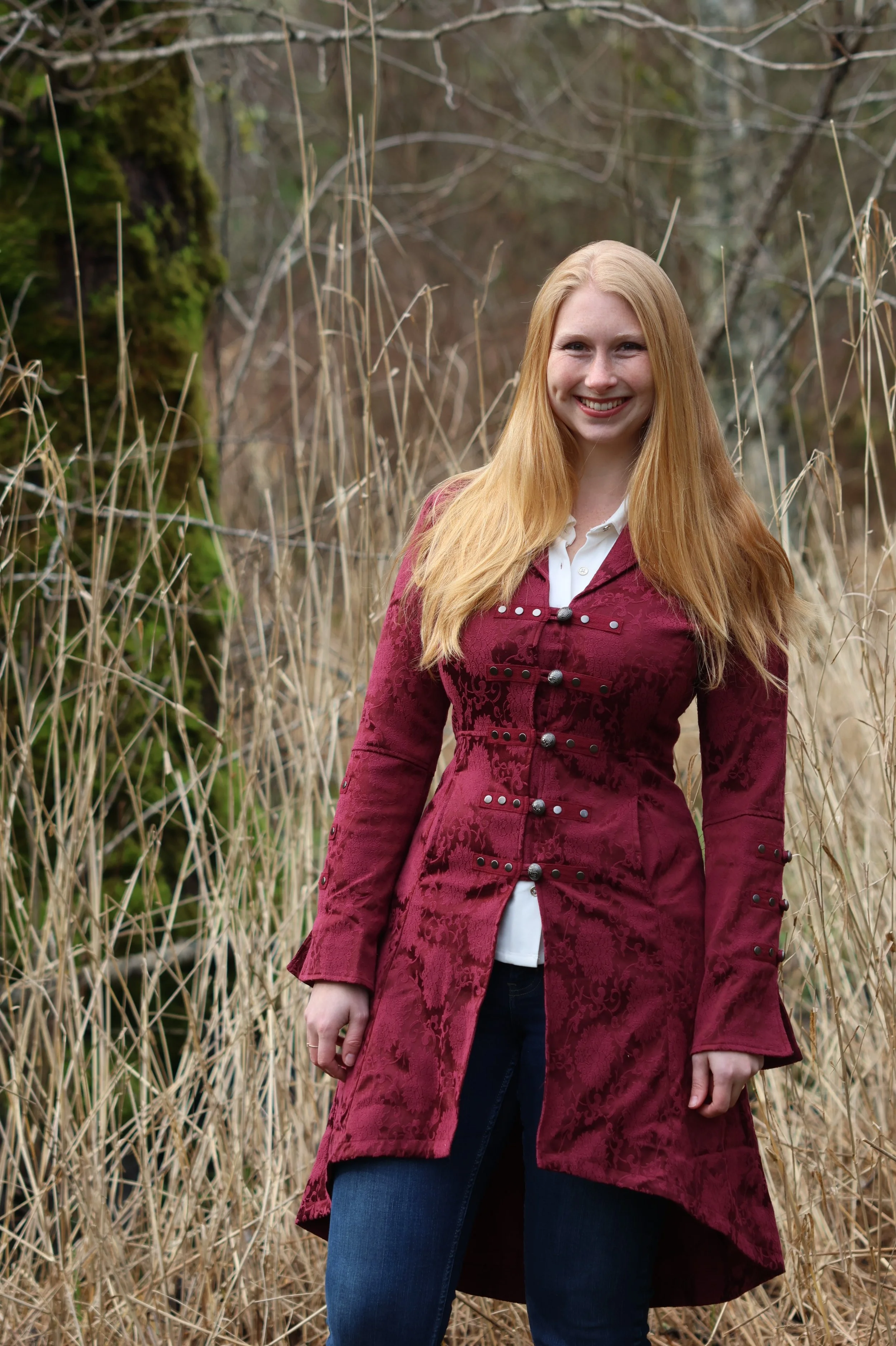 A woman with long red hair standing in a field of tall, dry grass with trees in the background, smiling at the camera.