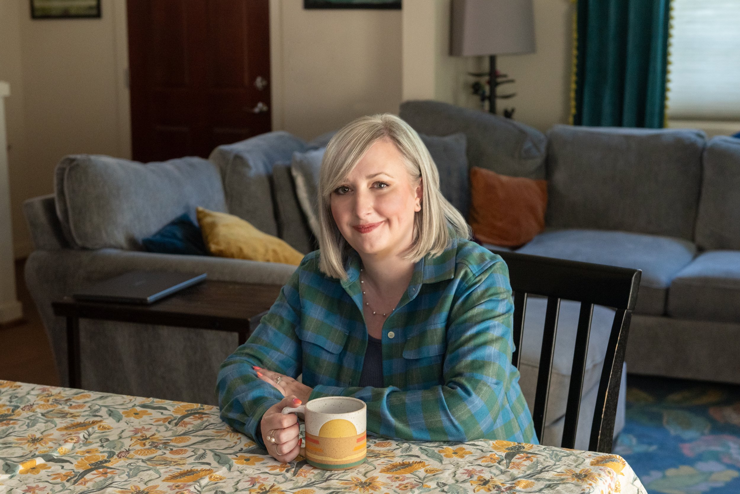Adrienne Scritsmier with blonde hair in a plaid shirt sitting at a table with a coffee mug, in a living room with gray couches, colorful pillows, a window with teal curtains, and a door in the background.