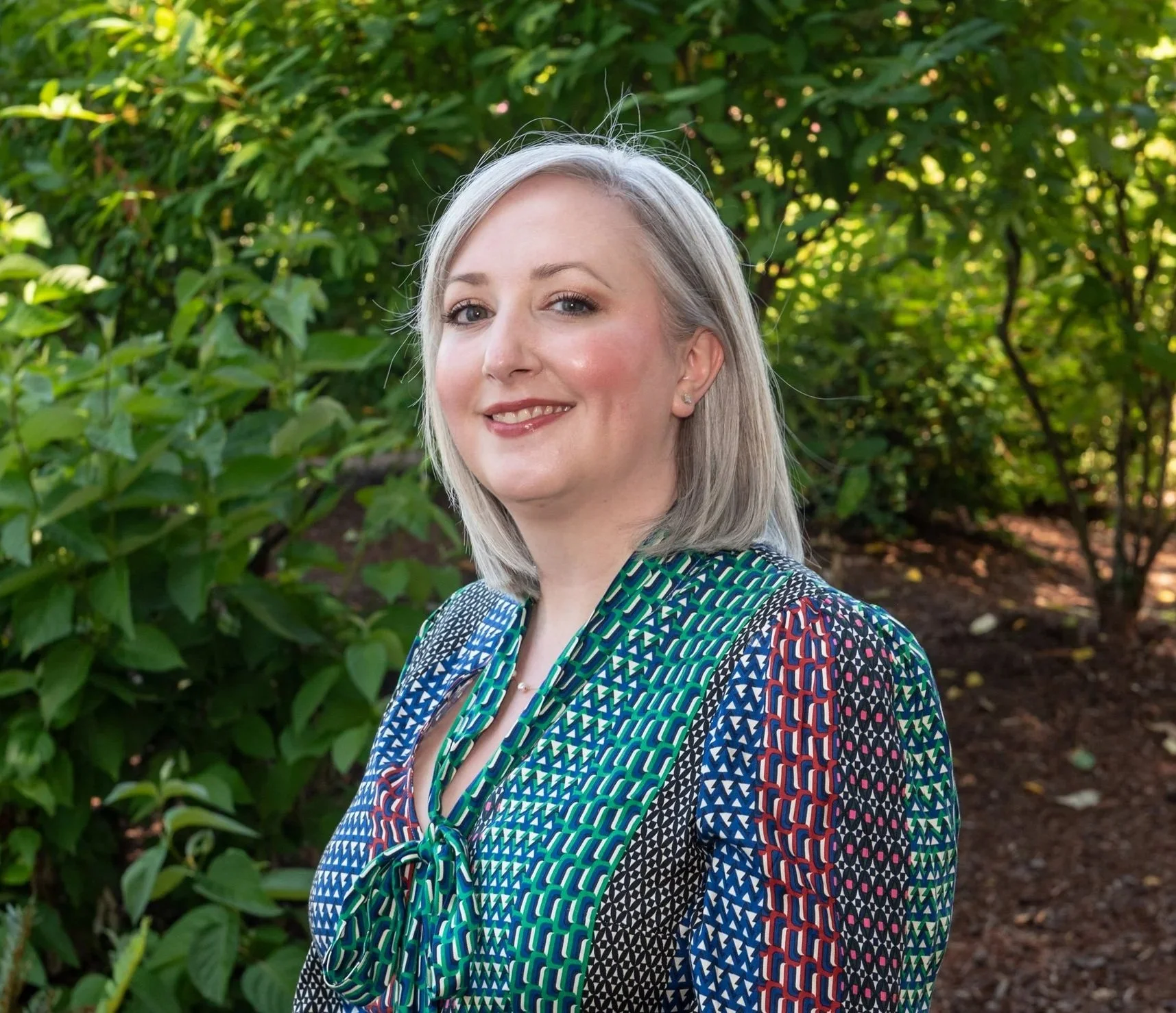 Portrait of a woman with blonde hair and light makeup, smiling, standing outdoors in front of green bushes, wearing a colorful patterned blouse.