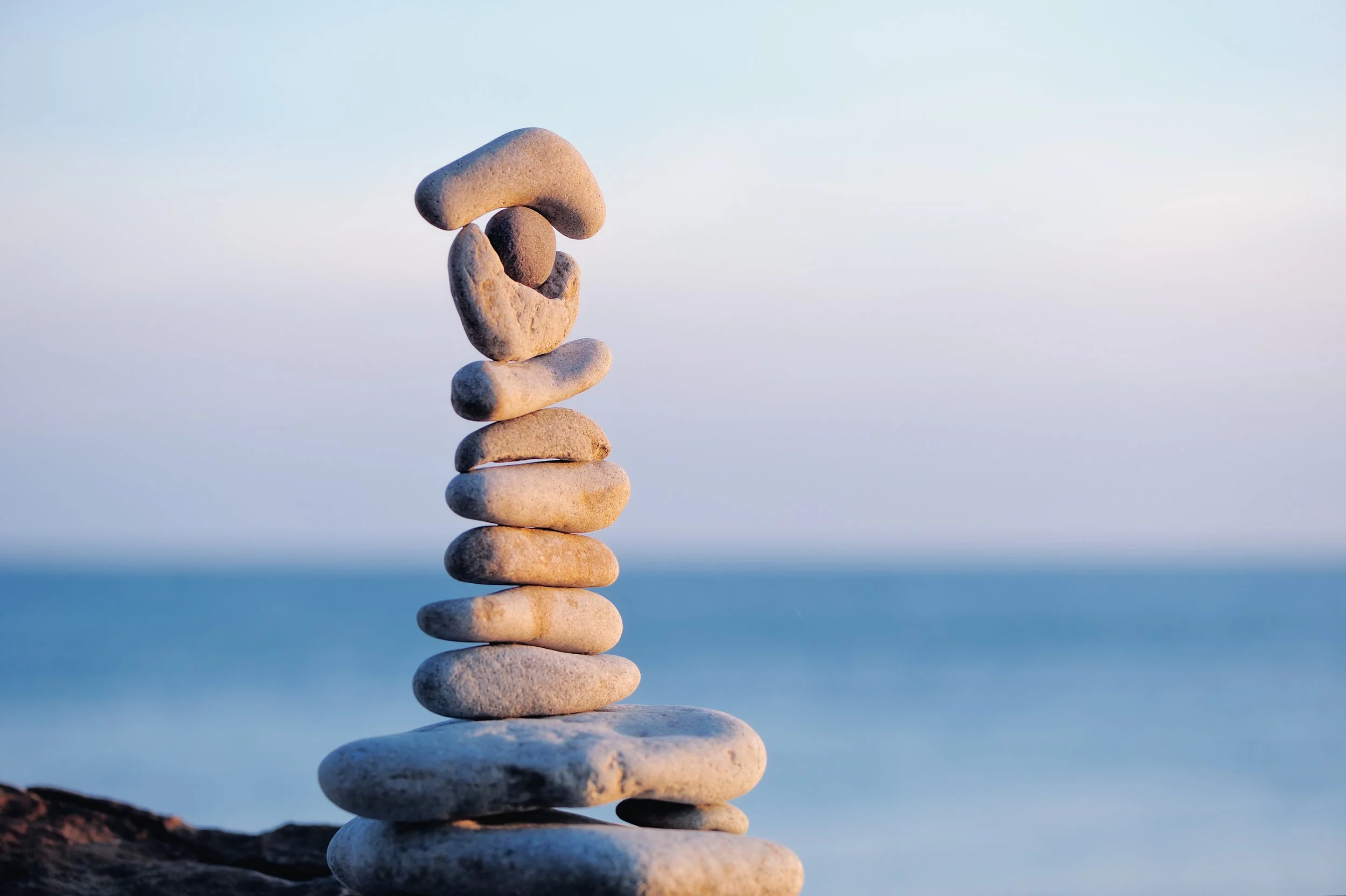 Stacked smooth stones balanced on each other near the water with a calm blue sky background.