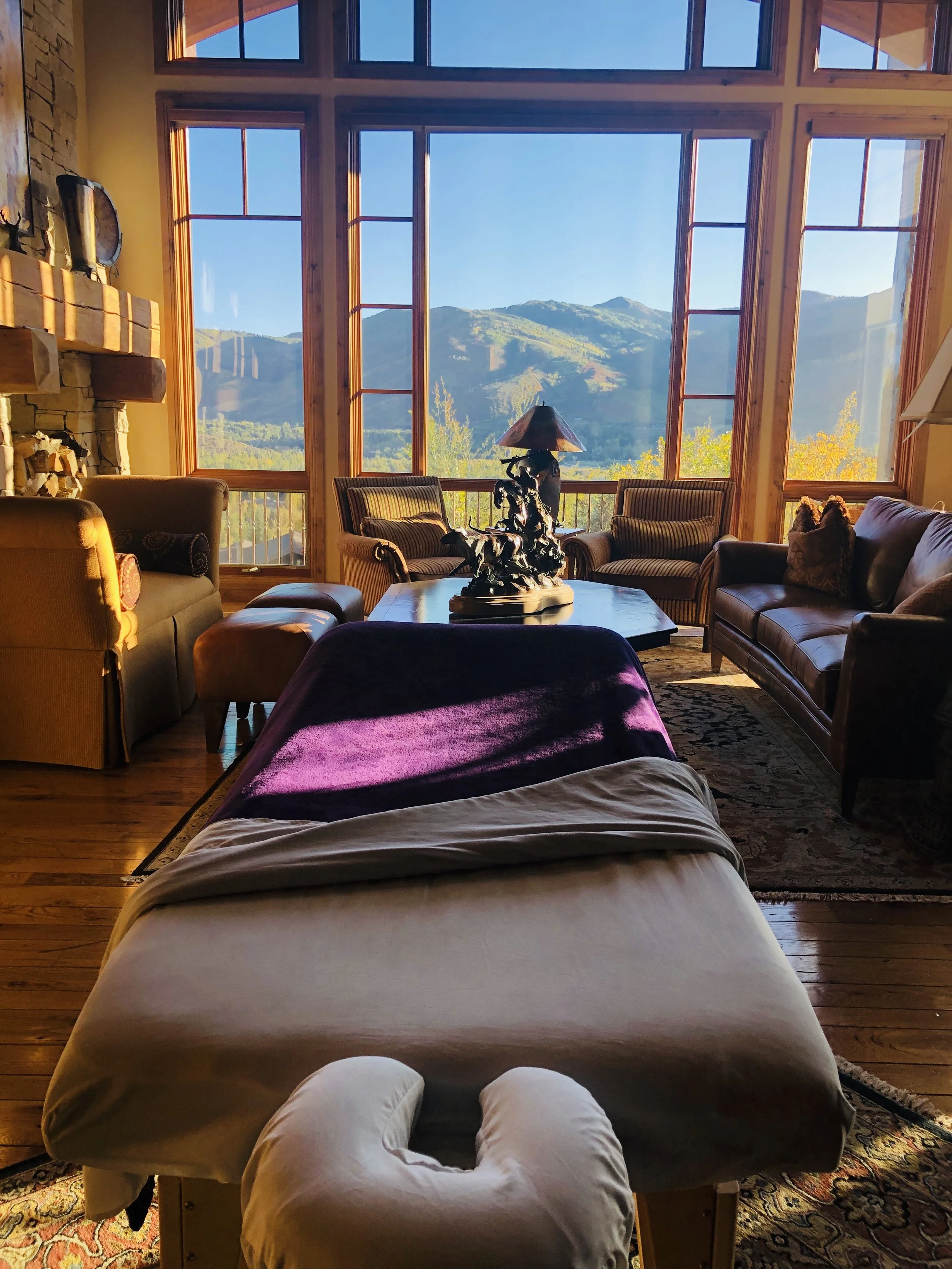 View of a cozy living room with large windows showing mountain scenery, a stone fireplace, a coffee table with a decorative lamp, and multiple chairs and sofas arranged for relaxation.
