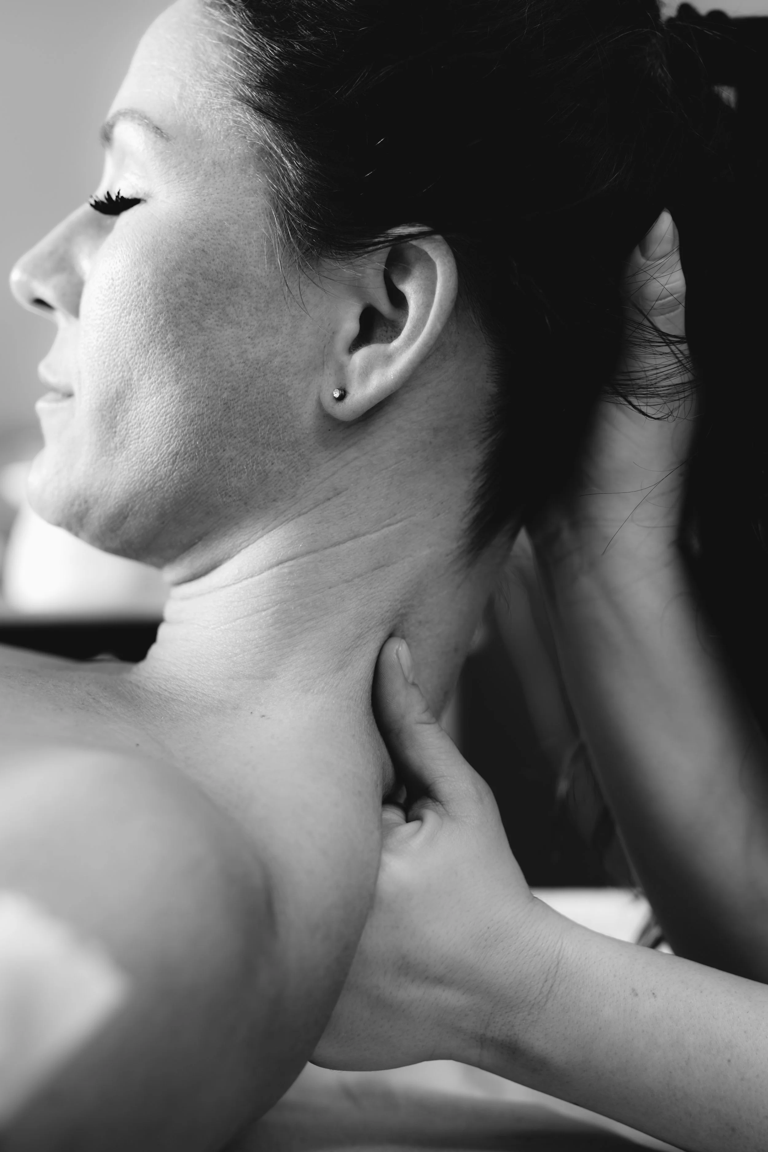 Black and white photo of a woman with closed eyes, dark hair, and a small earring, seen from the side, touching her neck with one hand.