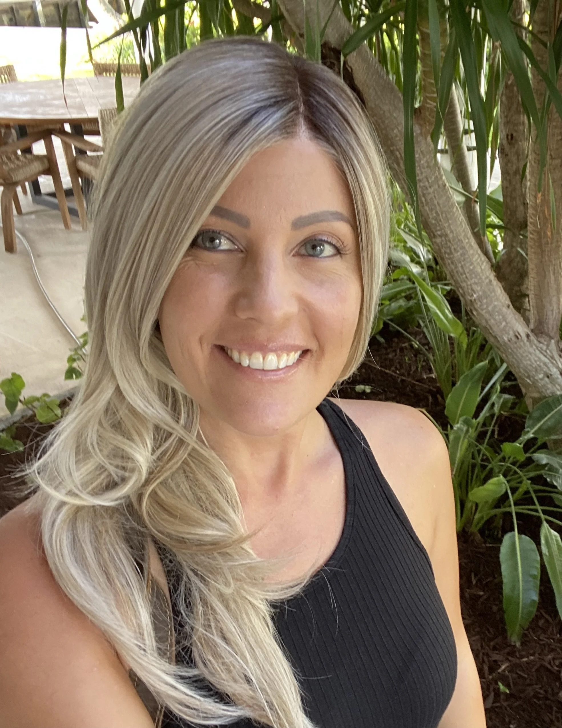 A woman with blonde hair, blue eyes, and a bright smile, wearing a black sleeveless top, standing outdoors near greenery and trees.