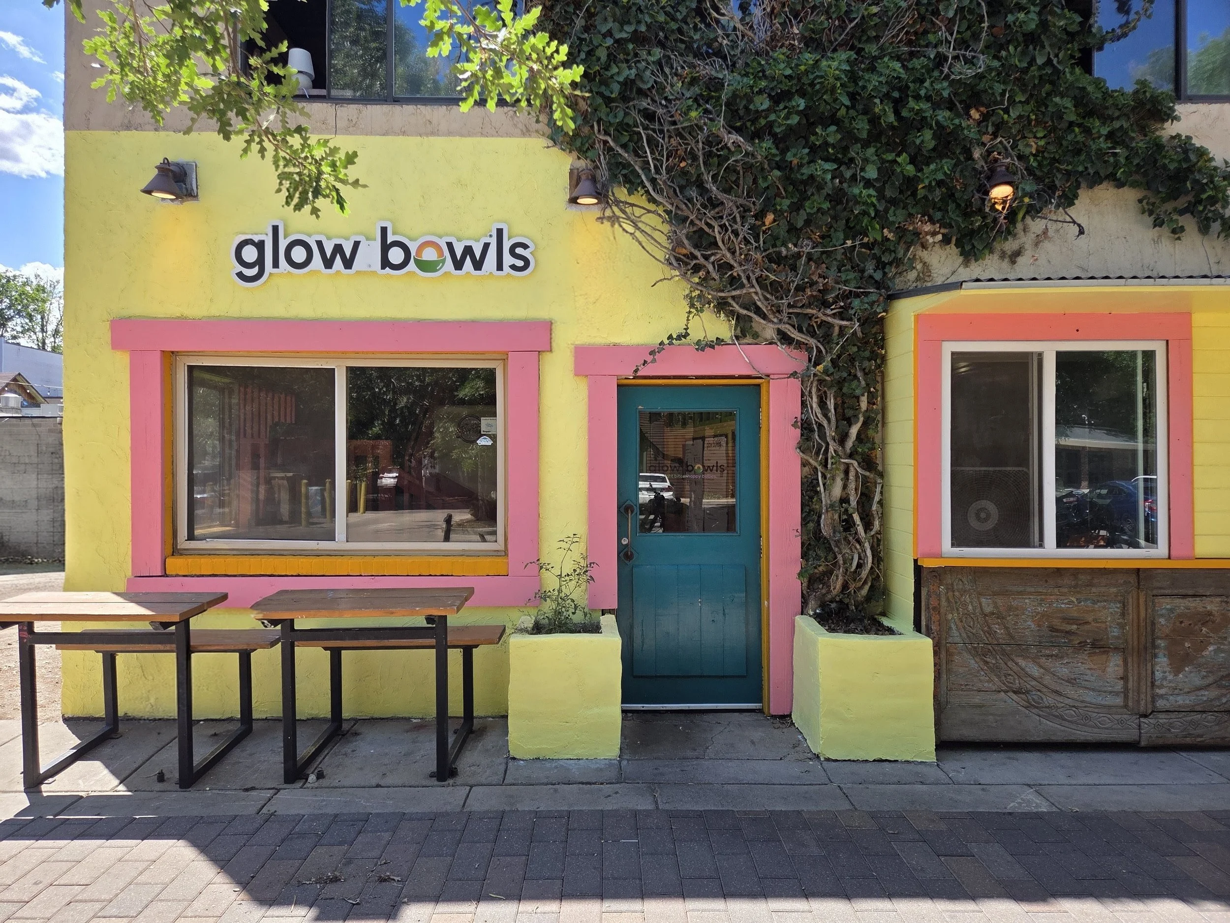 The exterior of a colorful building with a bright yellow facade, pink trim around the windows and door, and a green door. There is lush greenery with vines on the right side of the building and a large tree with green leaves on the left. The building has two large front windows and a sign that reads 'glow bowls' on the upper part of the front wall. In front of the building, there are metal tables with wooden tops on a paved sidewalk under a clear blue sky.