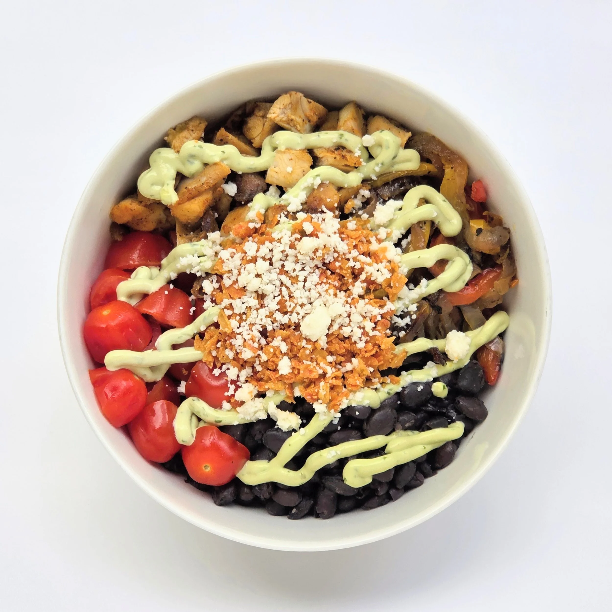 Close-up of a bowl filled with black beans, cherry tomatoes, chopped chicken, shredded cheese, and drizzled green sauce on top.