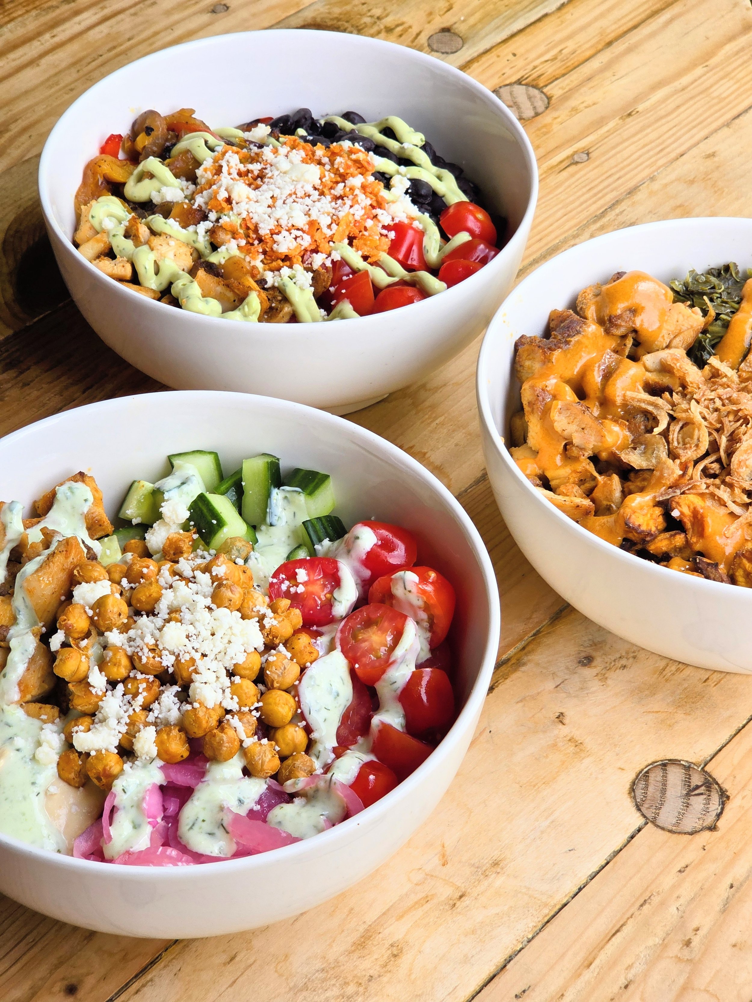 Three white bowls of colorful salads and toppings on a wooden table.