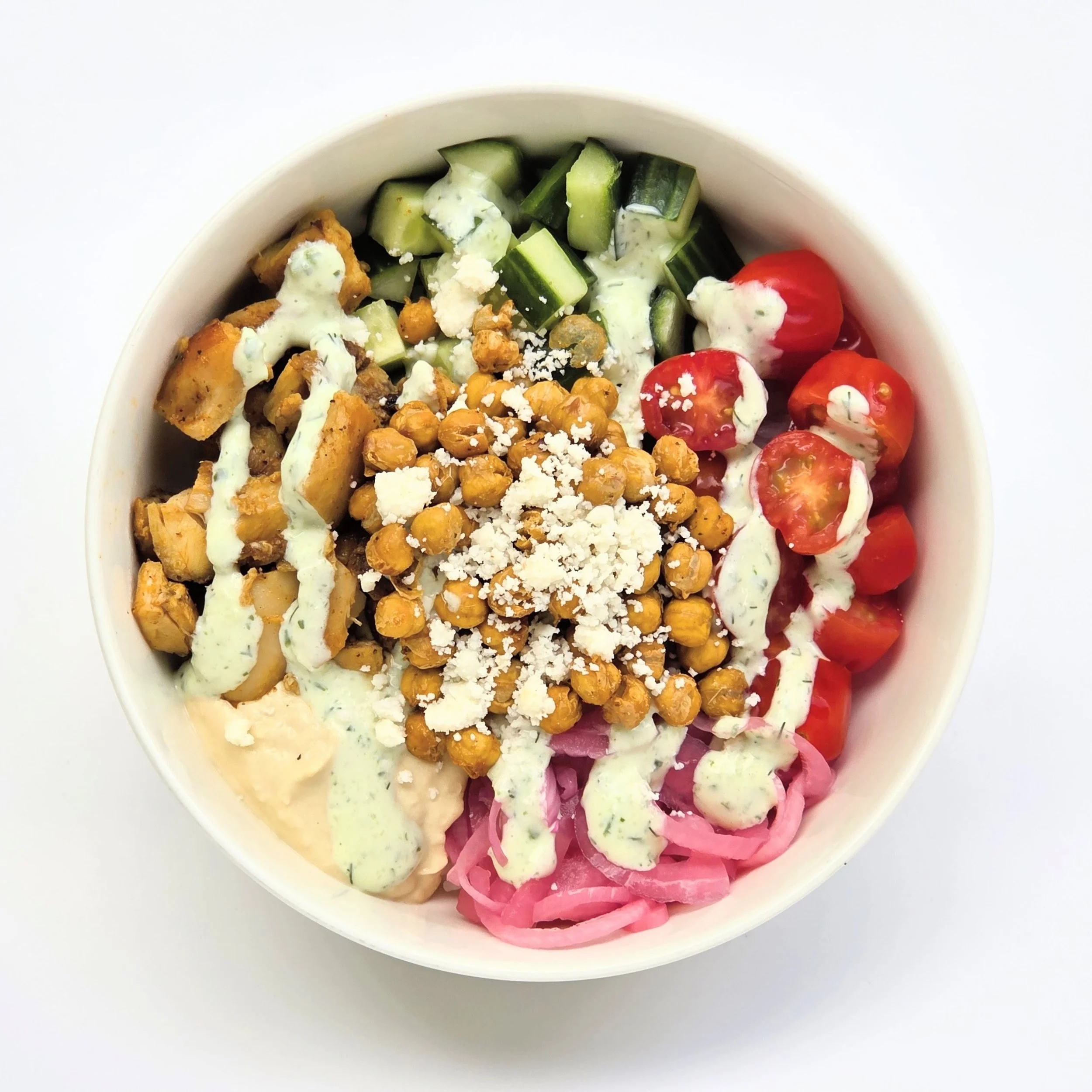 Close-up of a white bowl filled with a colorful salad containing cherry tomatoes, cucumbers, chickpeas, shredded cheese, chopped pink pickled onions, and a drizzle of creamy dressing.