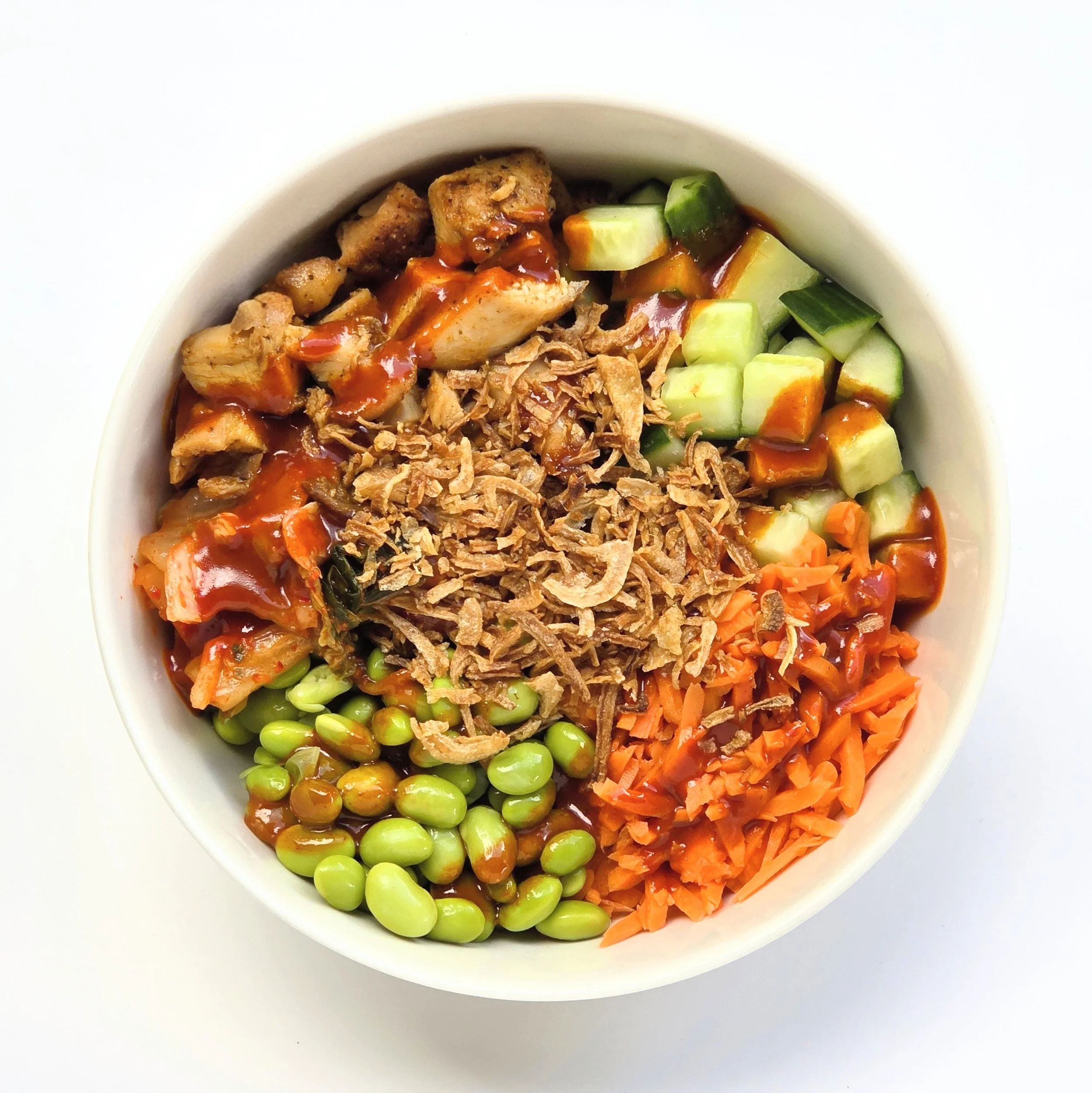A white bowl filled with diced cucumbers and shredded carrots on one side, shredded chicken with sauce on another, and green peas scattered throughout, all on a white surface.