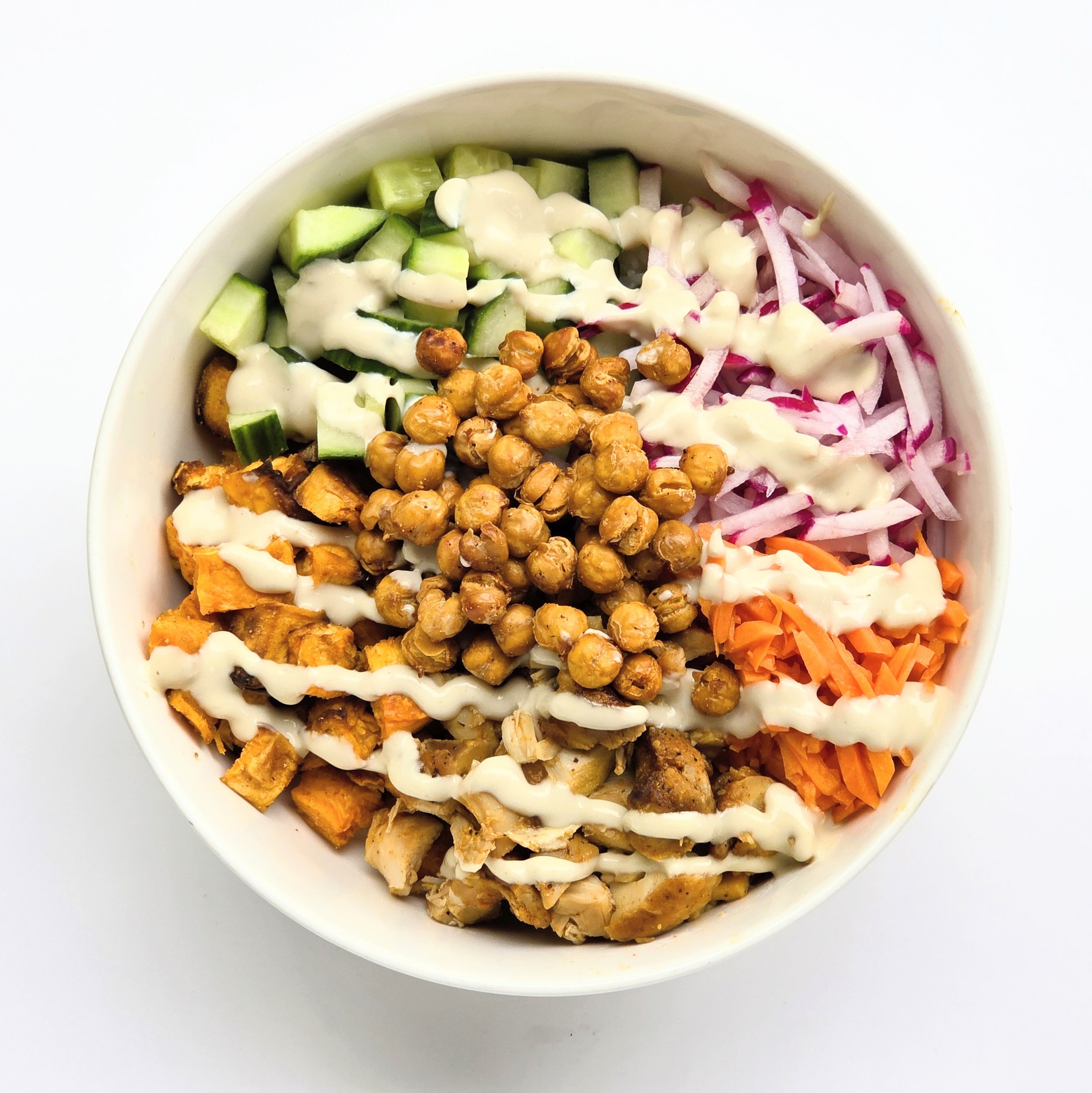 A white bowl filled with chopped cucumbers, shredded radishes, chickpeas, shredded carrots, chopped chicken, and drizzled with dressing on a white surface.