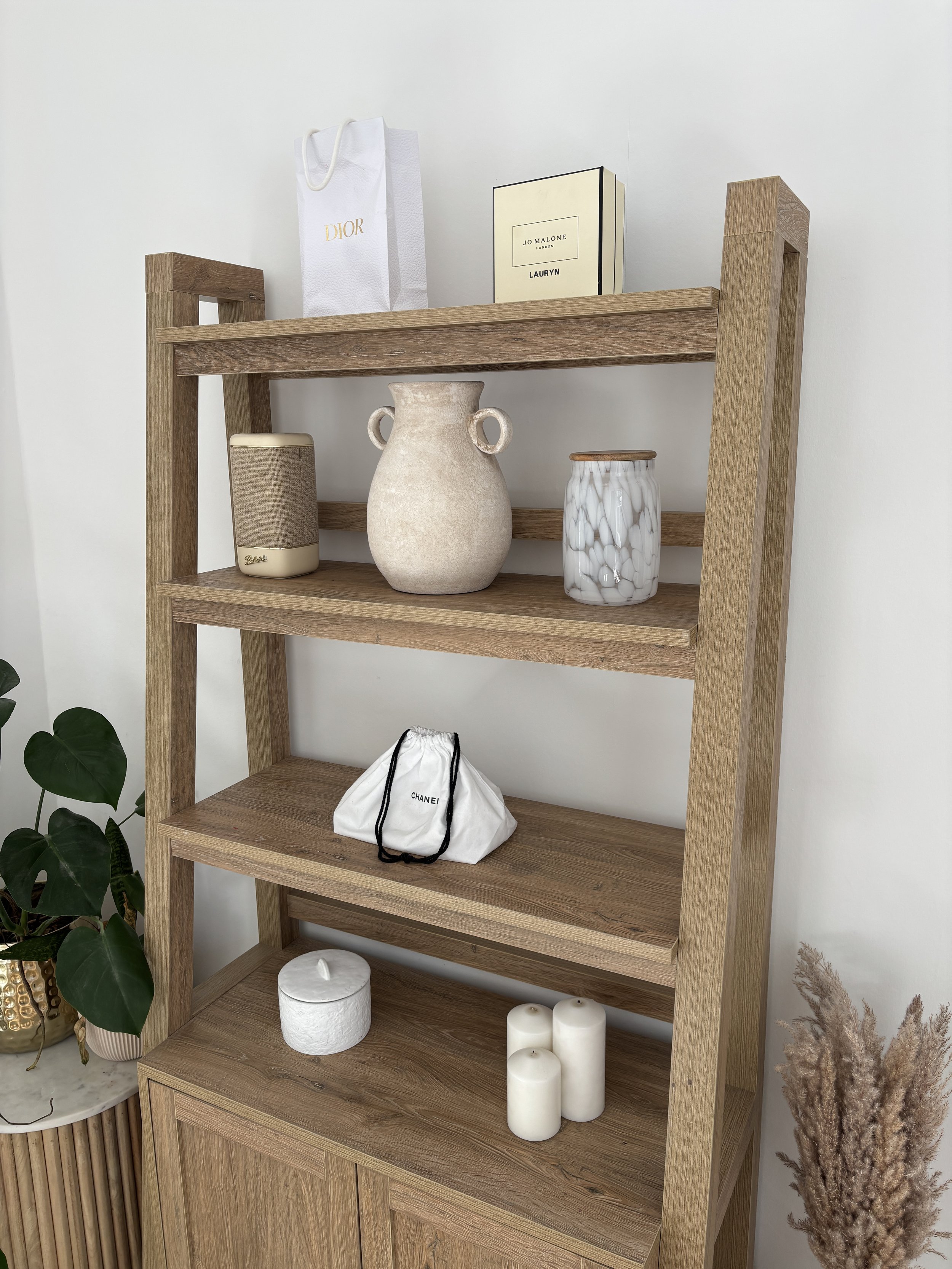 Wooden shelf with decorative items: a Dior shopping bag, Jo Malone box, a beige earthenware vase, a white and gray marbled jar, a white Chanel bag, white candles, a potted plant with large green leaves, and dried pampas grass.
