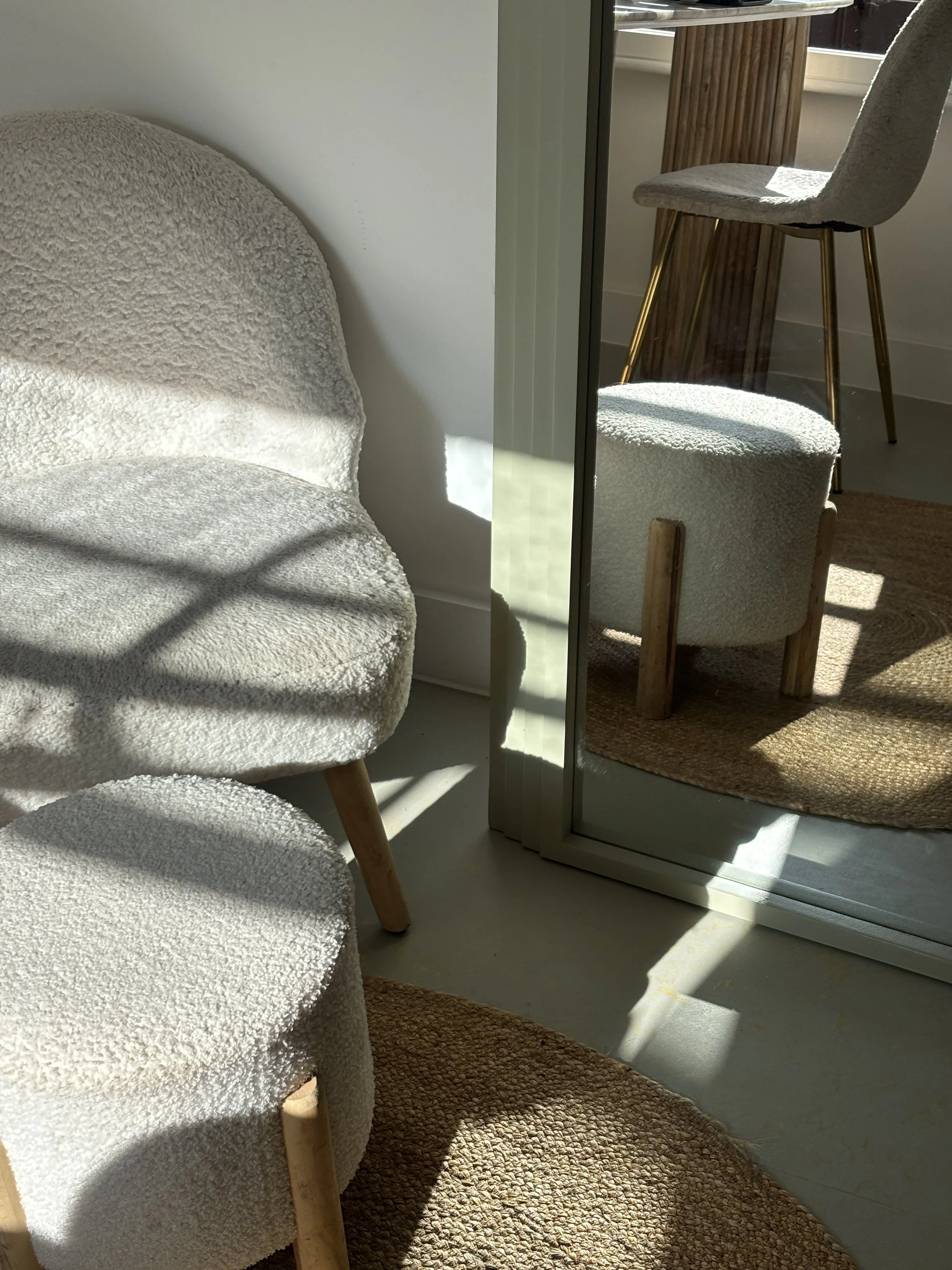 Close-up of a cozy corner of a room showing a plush beige armchair with wooden legs and a textured fabric, a matching round ottoman, a rustic brown area rug, and a mirror that reflects a wooden table with a beige upholstered chair.