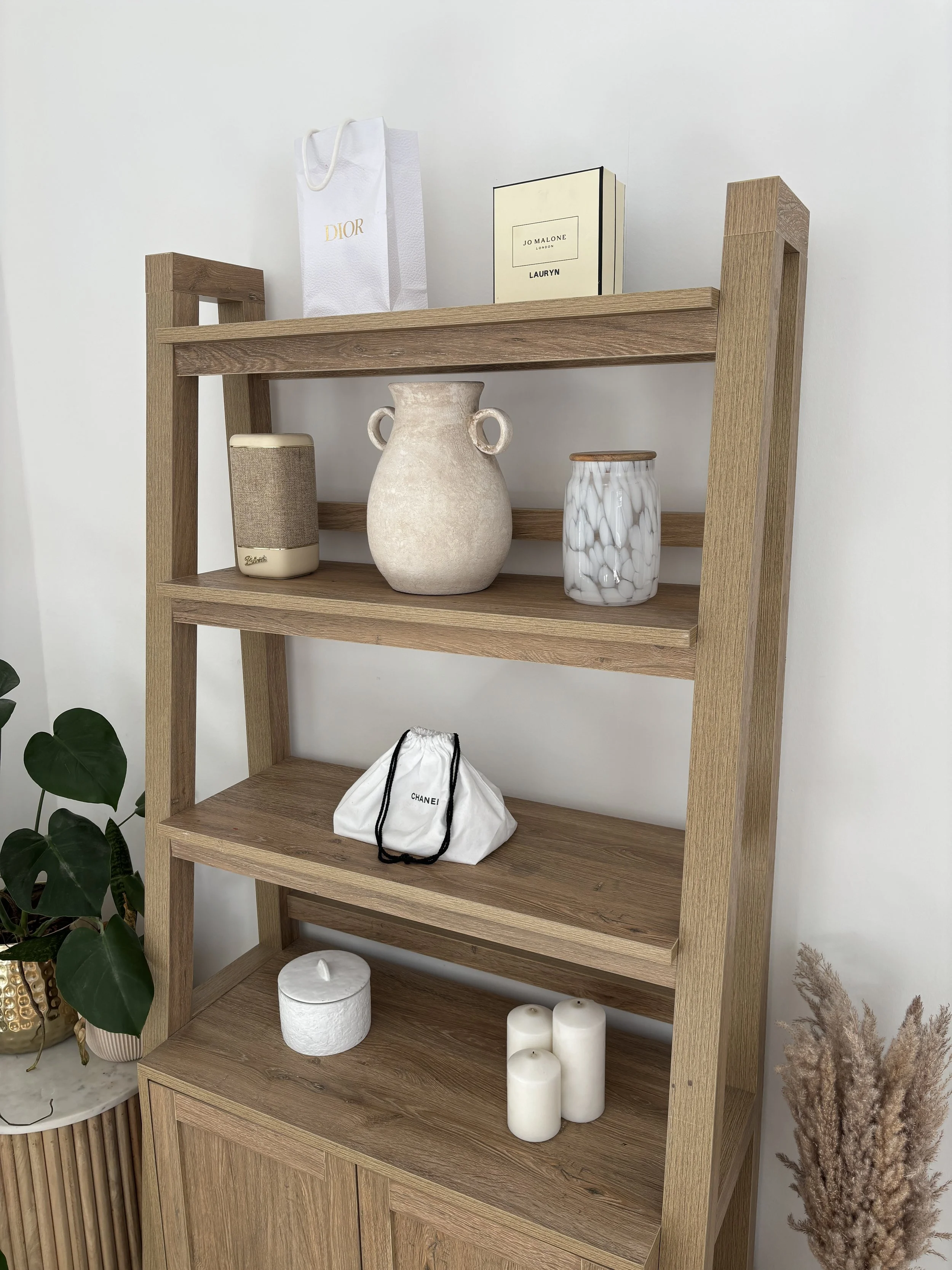 Wooden bookshelf displaying various decorative items such as candles, a ceramic vase, a small box, and a shopping bag, with a potted plant and pampas grass nearby.