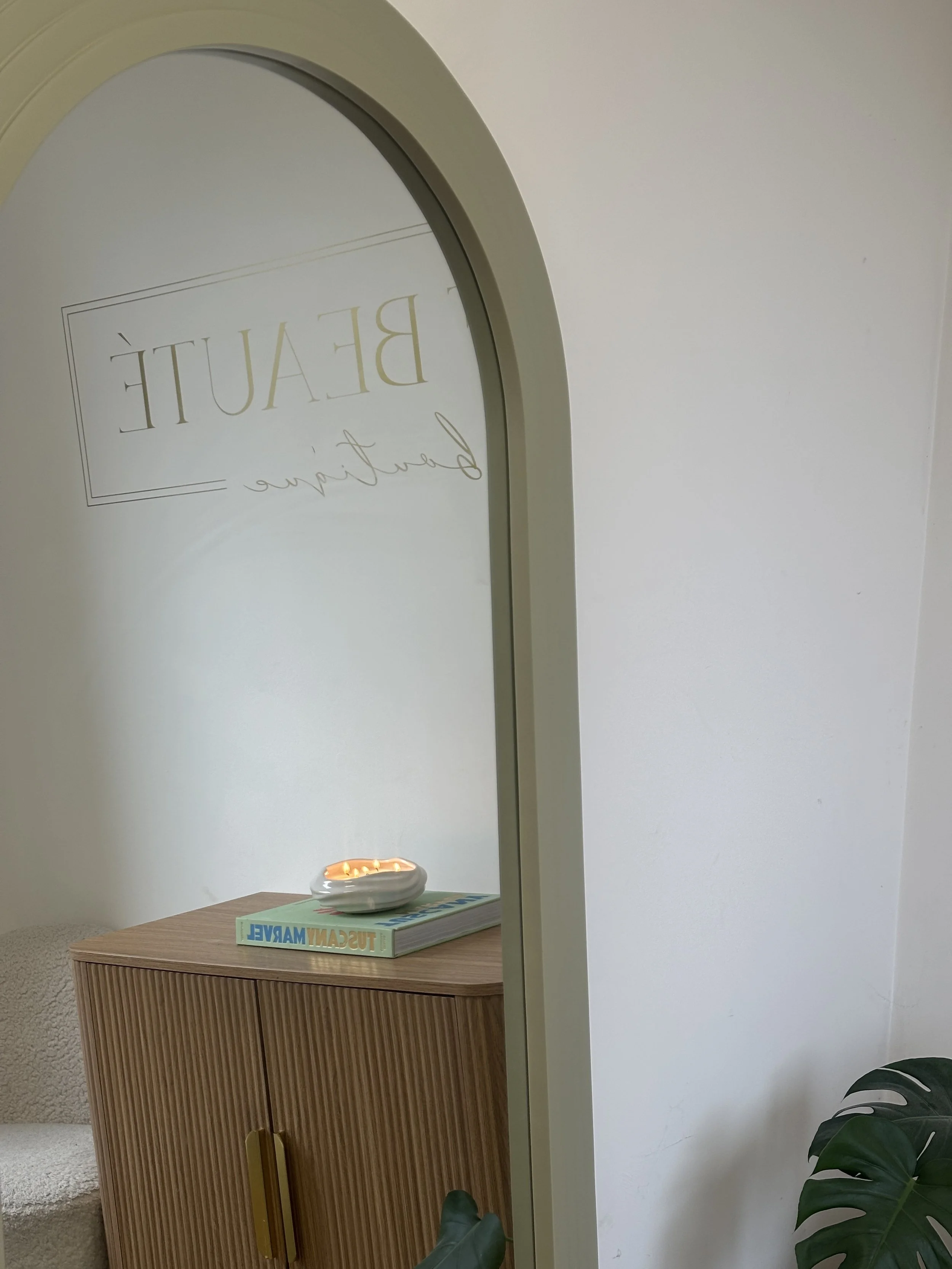 A mirror with a white frame reflecting the words 'BEAUTE' and 'beautiful' in script, mounted on a white wall. Below the mirror, a small wooden cabinet with a book titled 'TUSCAN MARKET' and a white shell-shaped object on top. To the right, part of a 