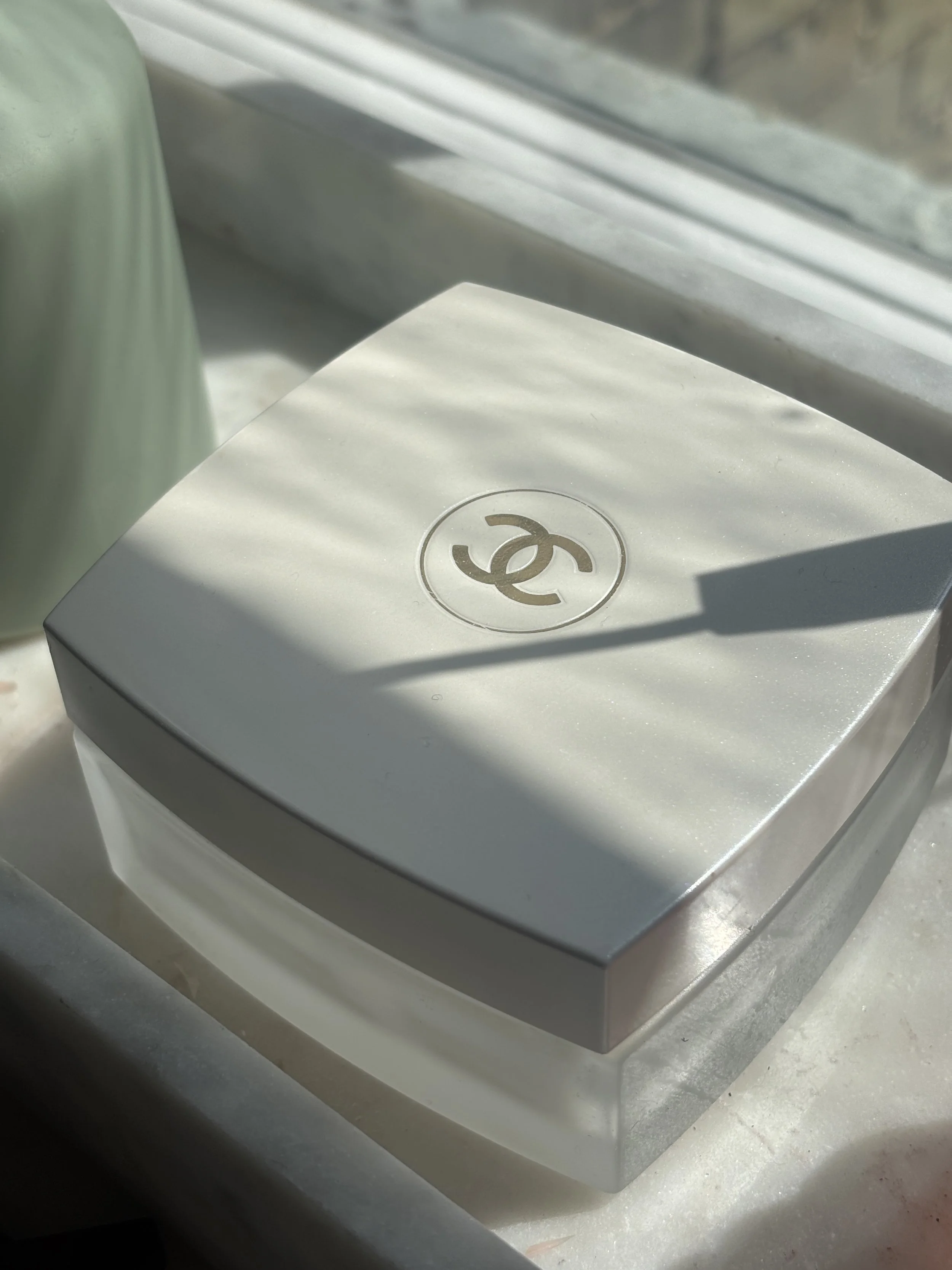A white box with a black Chanel logo on the top, placed on a flat surface. The box is partly covered in shadows, with some sunlight illuminating part of it.