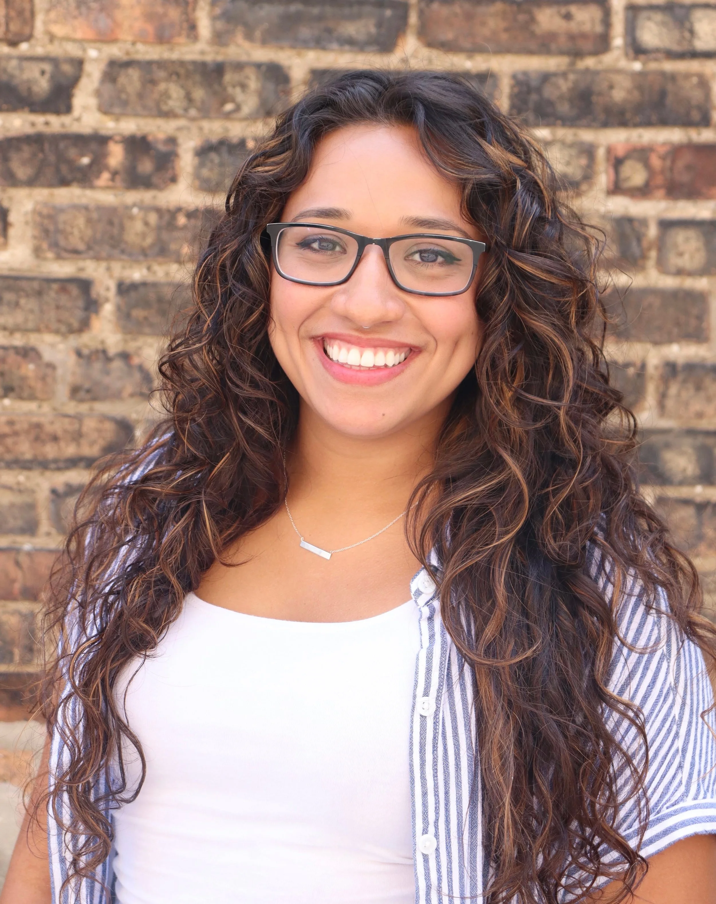 Close-up of a woman with curly dark hair, glasses, and a nose piercing, smiling in front of a brick wall.
