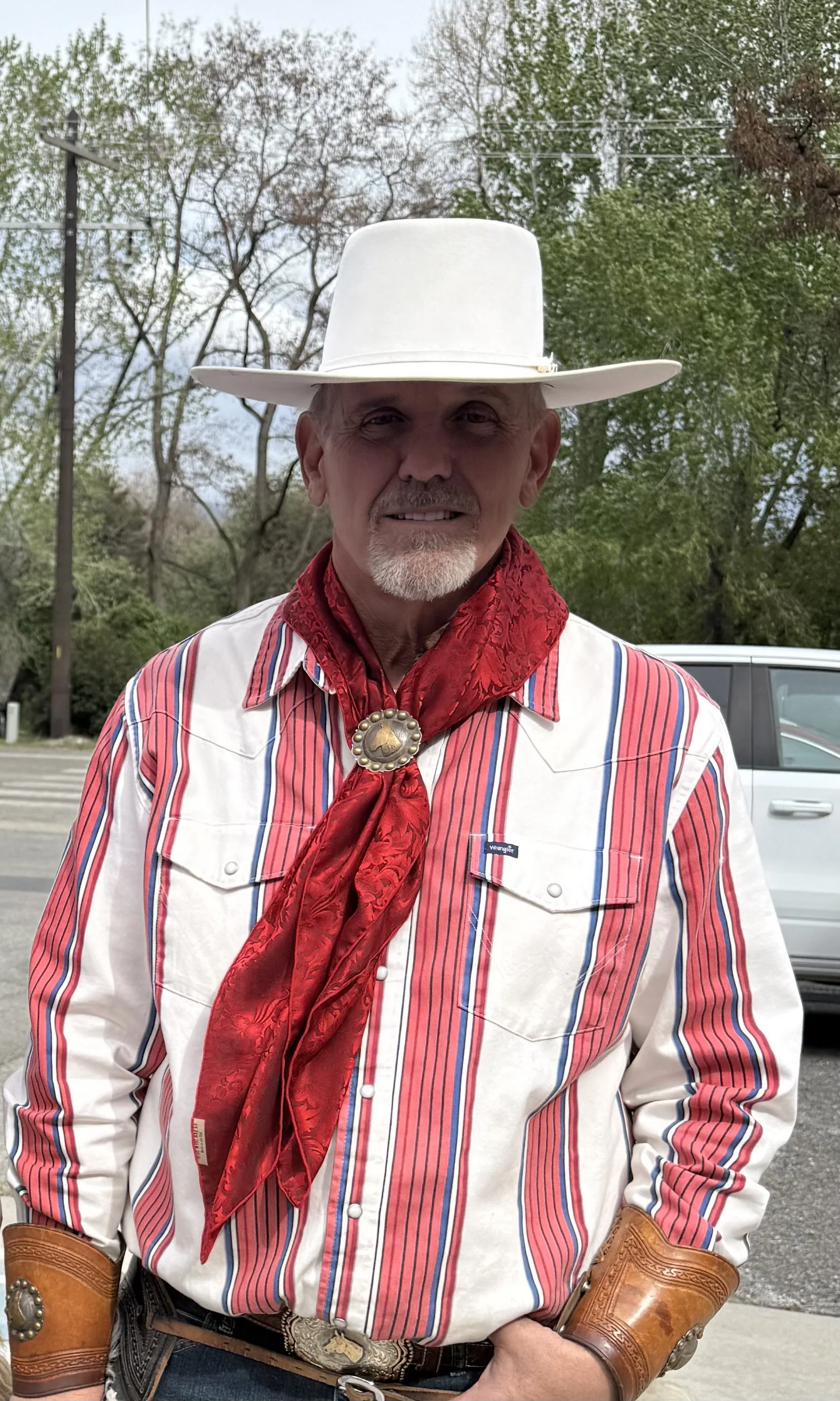 A man dressed in Western cowboy attire, including a white cowboy hat, red bandana with a silver concho, striped shirt, leather cuffs, and a belt with a decorative buckle, standing outdoors in front of a parking lot and trees.