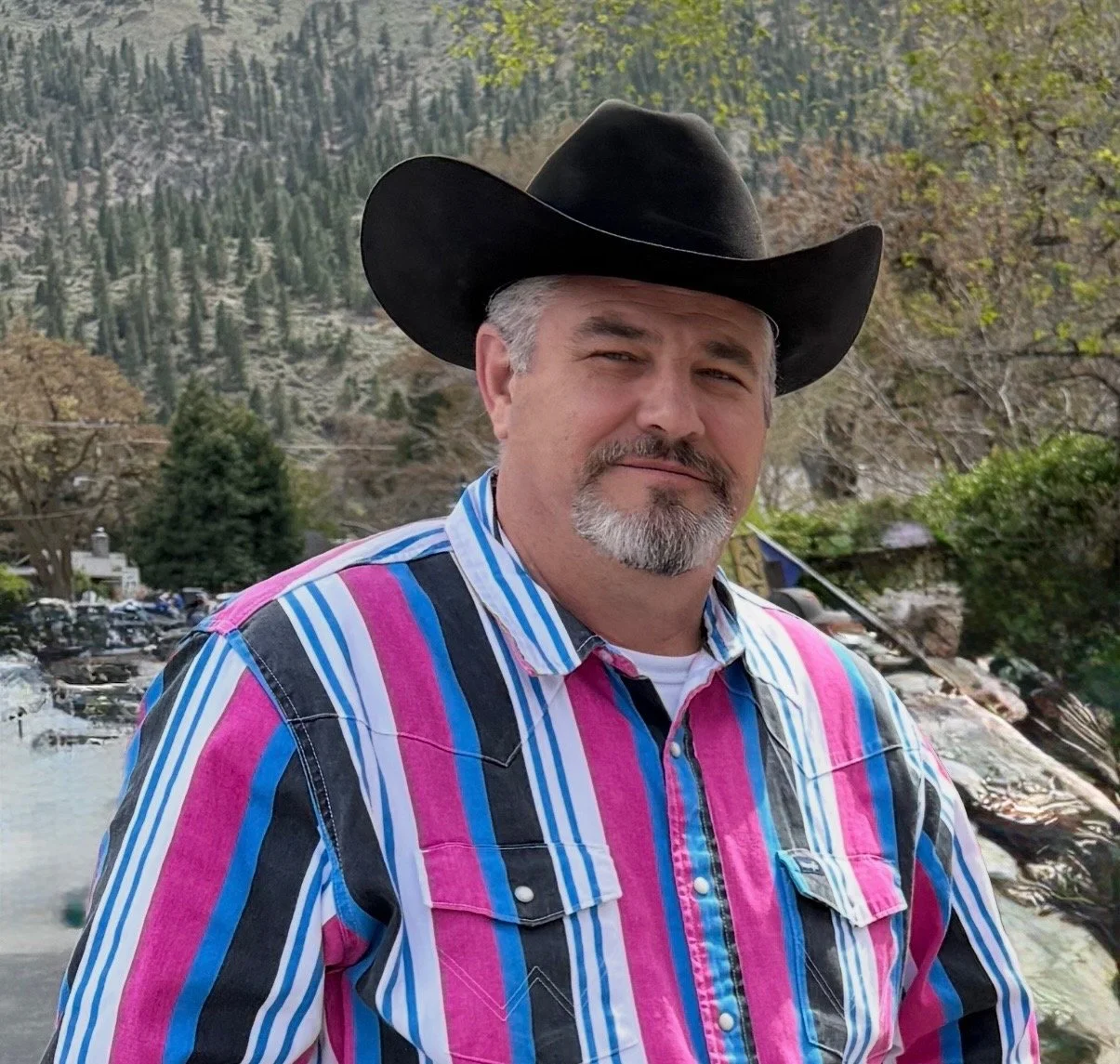 A man wearing a black cowboy hat and a colorful striped shirt standing outdoors with a mountain landscape, trees, and a river in the background.