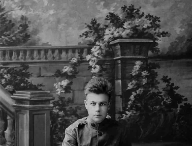 A young boy with short hair in a collared shirt sitting outdoors in front of a painted garden scene with trees, stone railings, and a large planter.