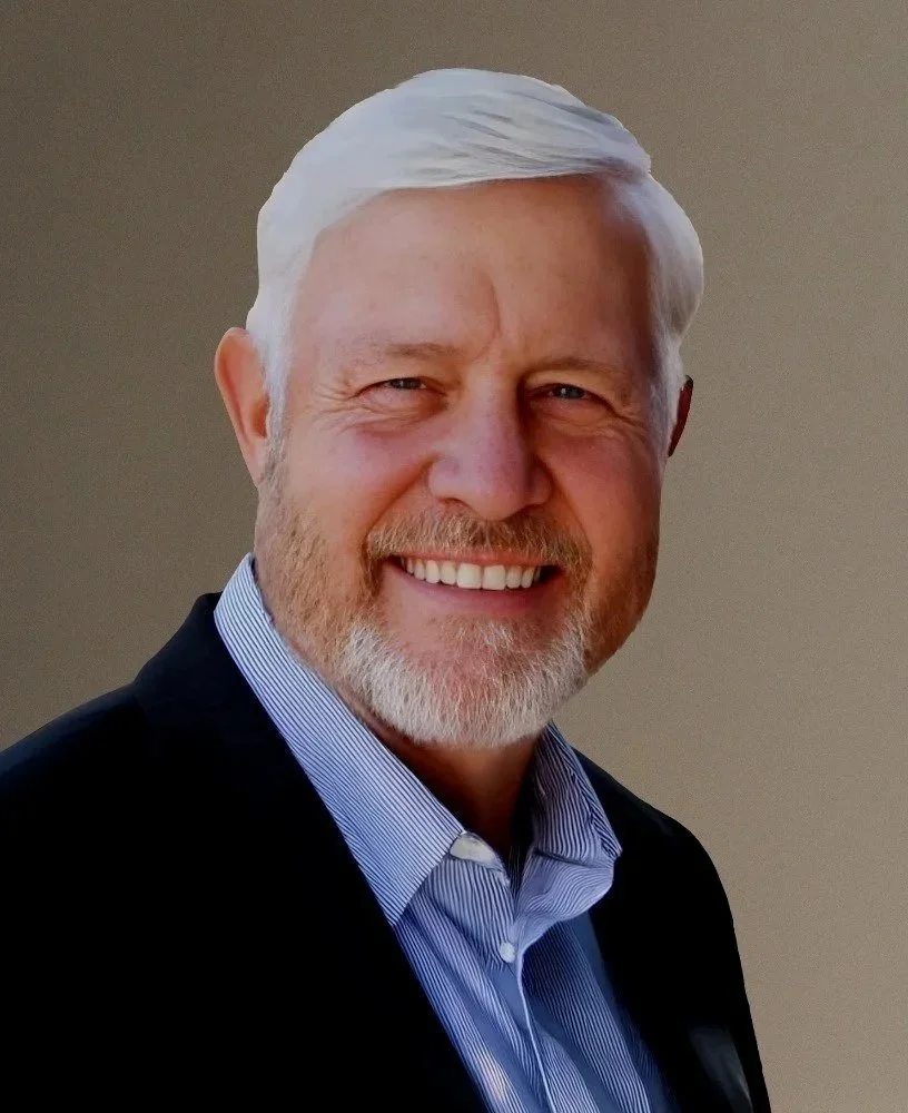 A smiling middle-aged man with white hair and a beard, wearing a dark suit jacket and a light blue collared shirt, against a neutral background.
