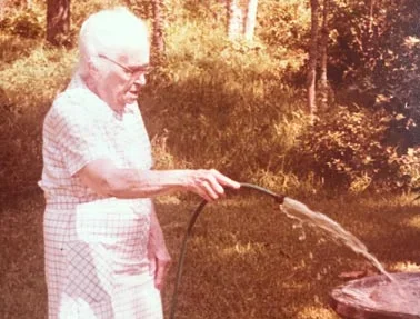 Elderly woman watering a garden or plants with a hose outdoors during daytime.