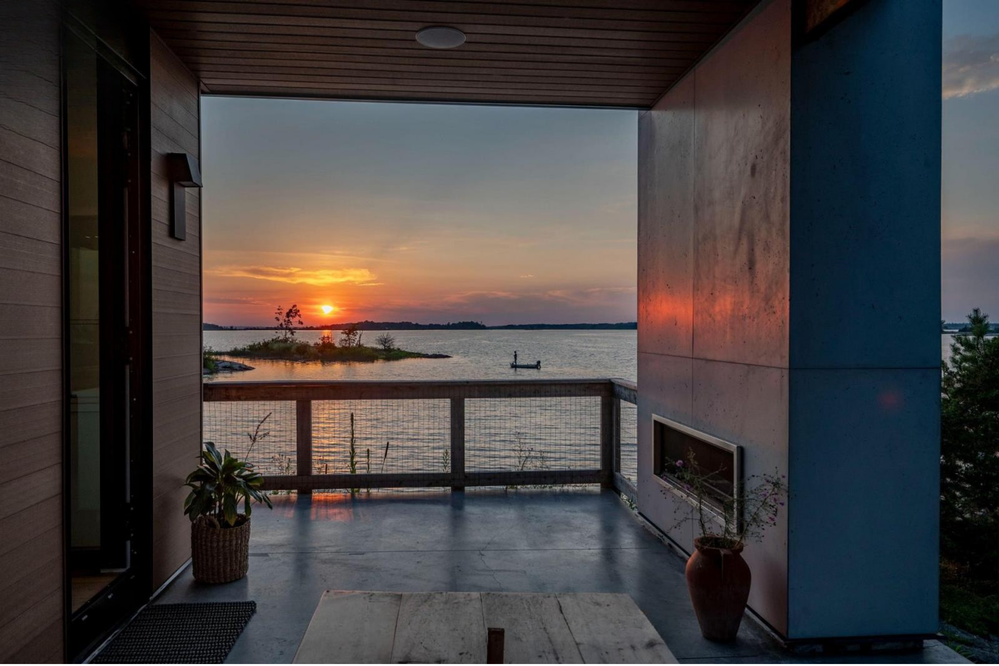 View of a sunset over a body of water seen from a balcony with a tiled floor, wooden walls, and a concrete wall; there are potted plants and a small wooden table on the balcony.