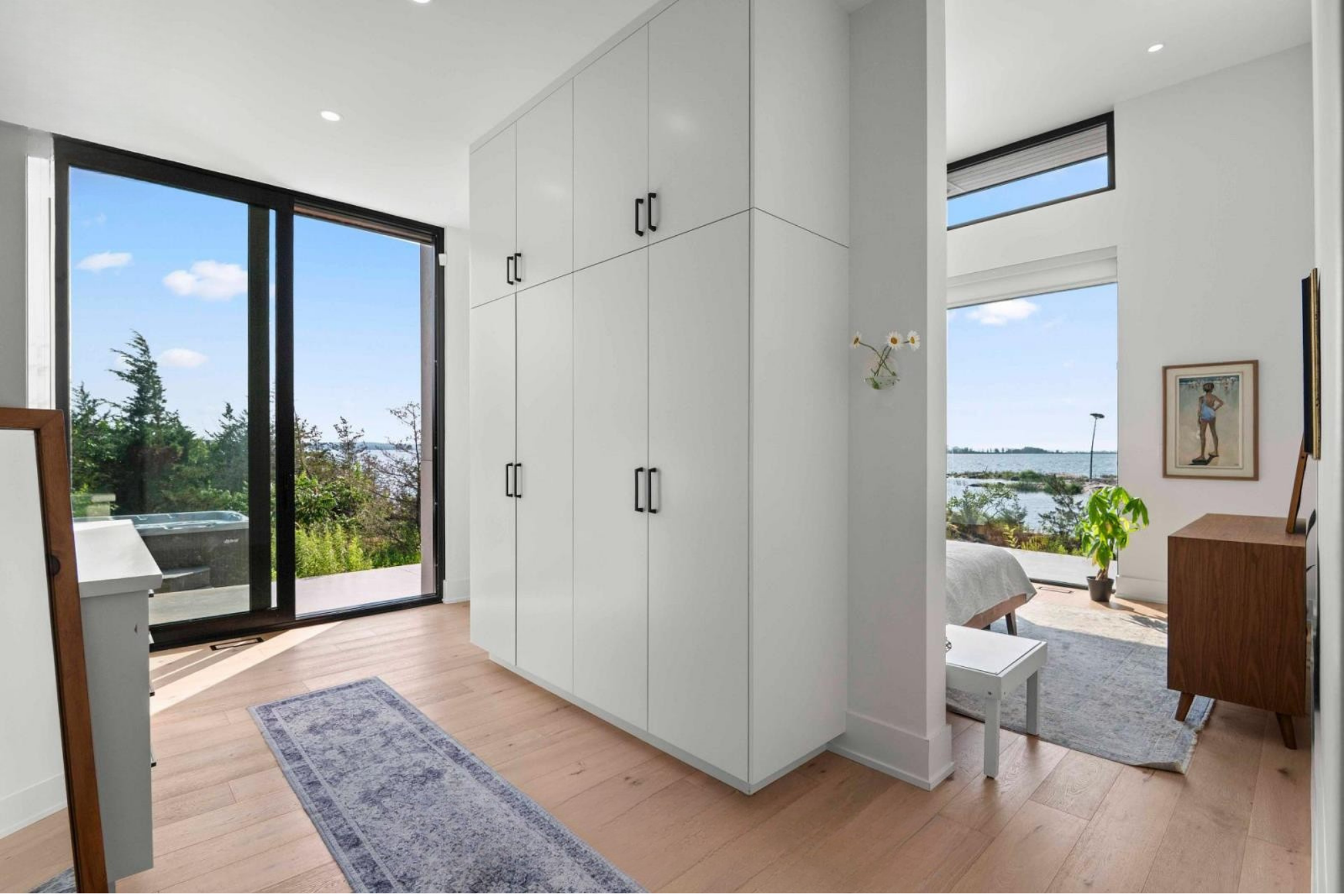 Bright modern living space with white walls, large windows showing a view of the water, a framed artwork, a potted plant, a small white table, and a white enclosed closet.