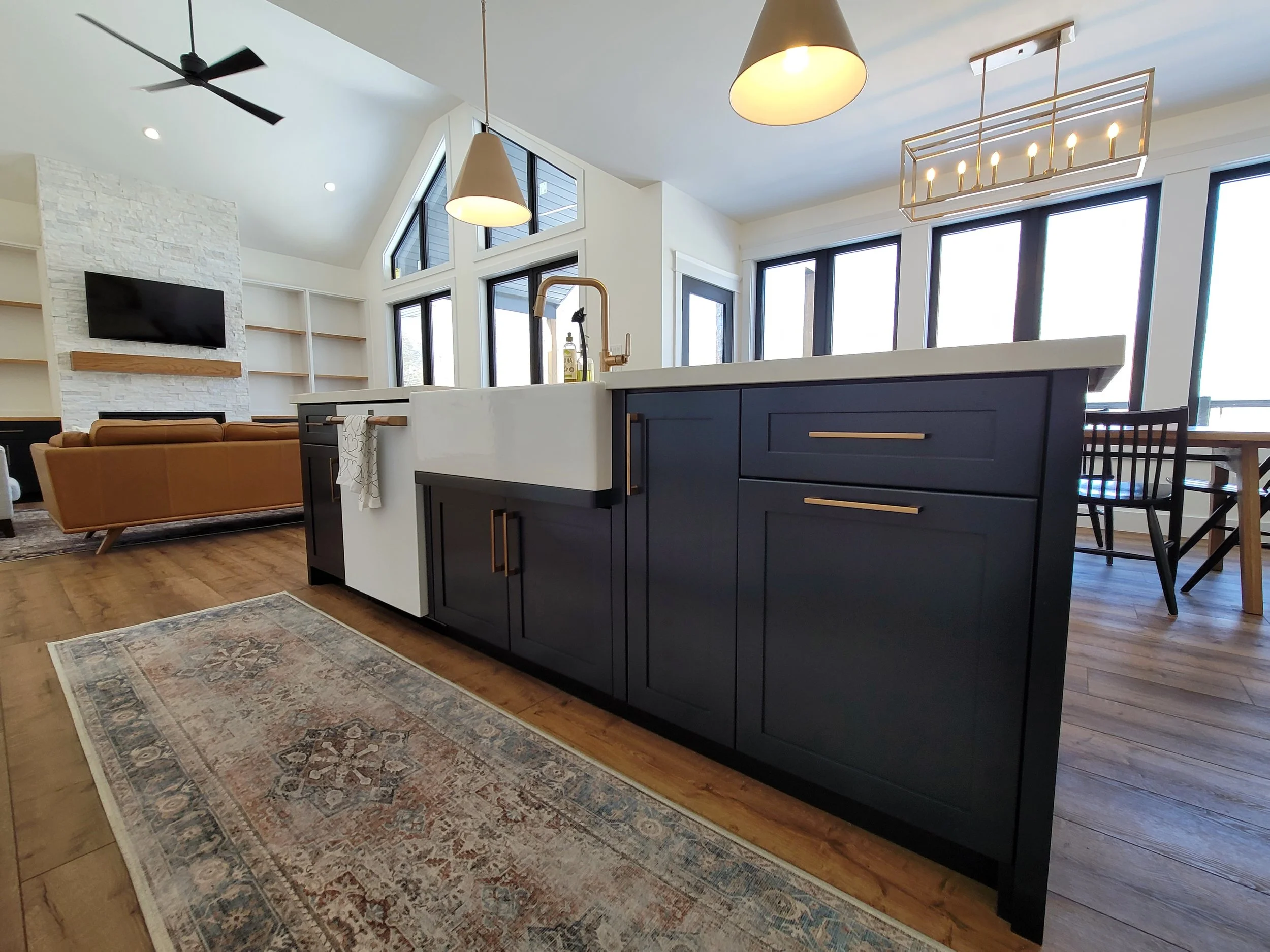 Open-concept living room and kitchen area with large windows, a black kitchen island, a tan sofa, wall-mounted TV, modern lighting fixtures, and a patterned rug.