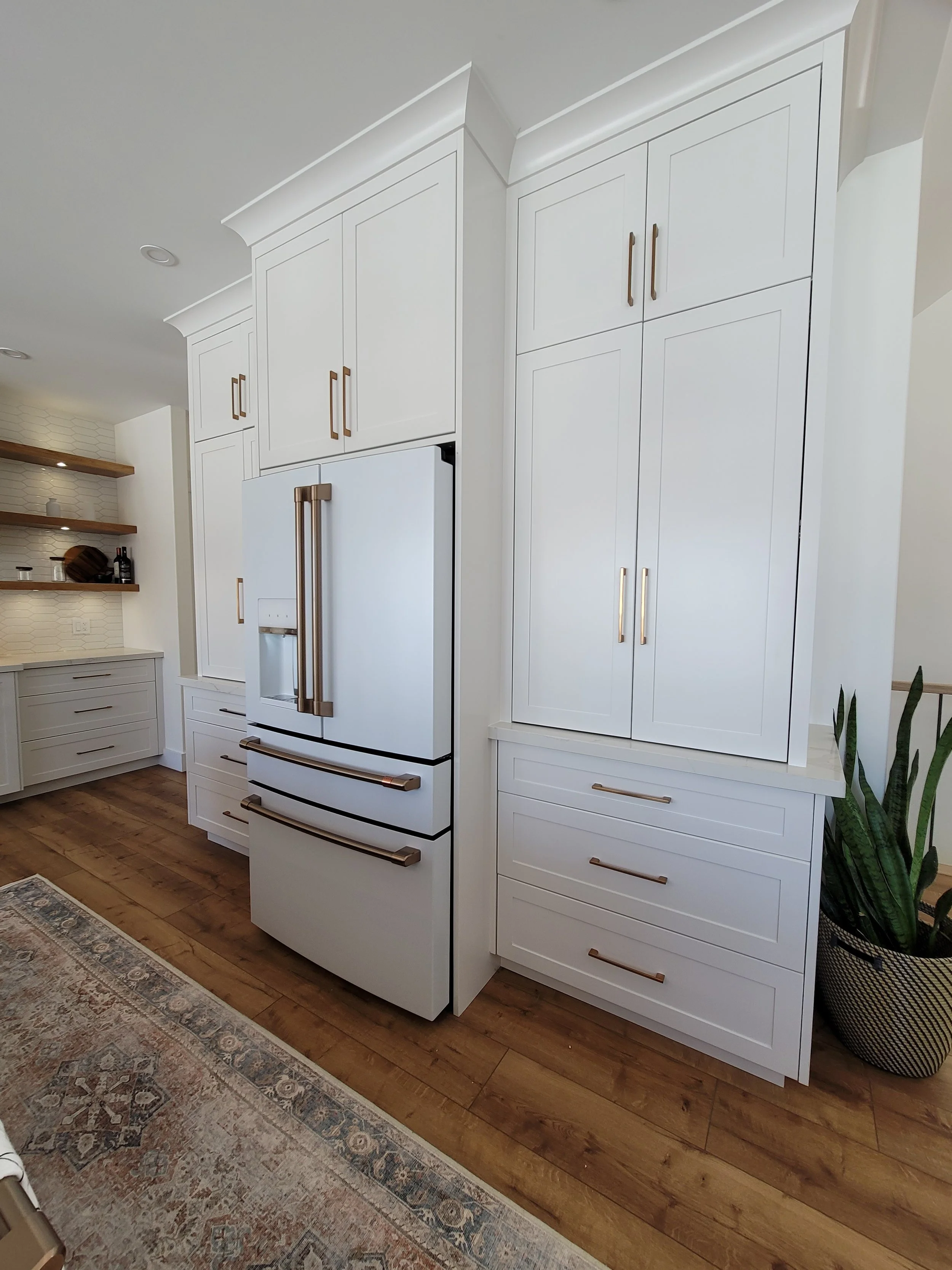 White kitchen cabinetry with gold handles, a large refrigerator, and wooden flooring. Part of a kitchen with open shelves and a patterned rug is visible.
