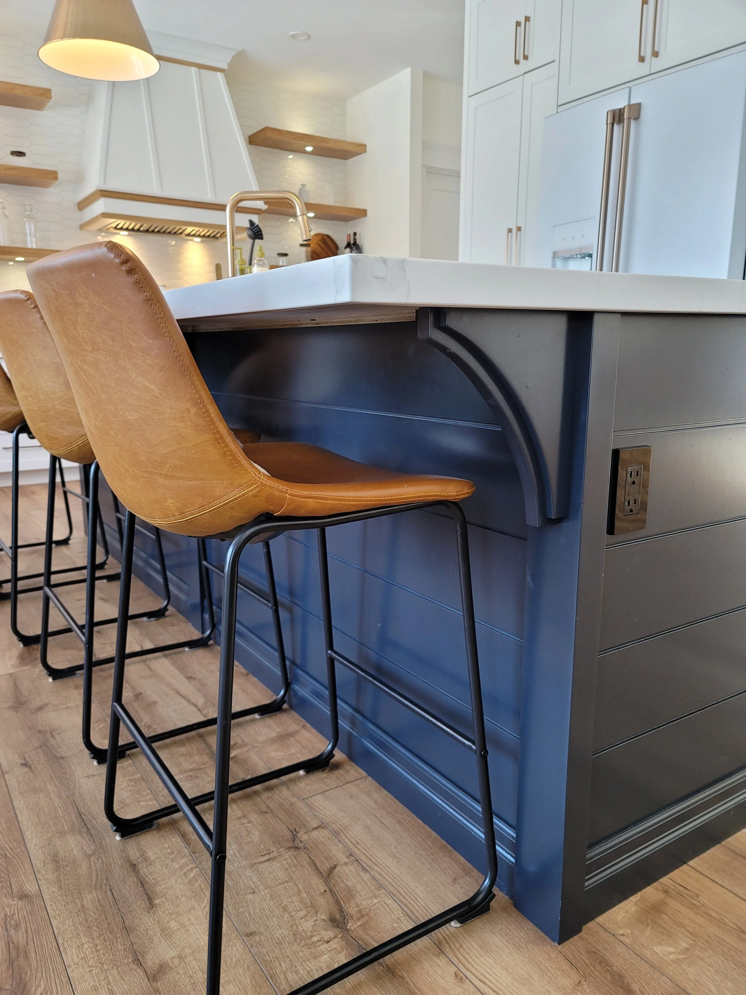 Contemporary kitchen with white cabinets, a large island with dark blue base and white marble countertop, brown leather barstools, wooden open shelves, and a wooden-floored kitchen area.