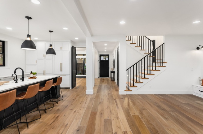 Modern open-concept kitchen and living area with hardwood floors, white cabinets, black kitchen fixtures, and a staircase with wooden steps and black railing.