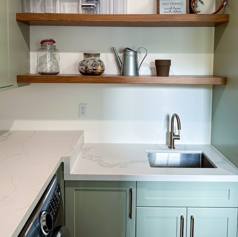 Kitchen with white marble countertop, stainless steel sink, green cabinets with vertical handles, open wooden shelves with jars and watering can, and a wall outlet on the backsplash.
