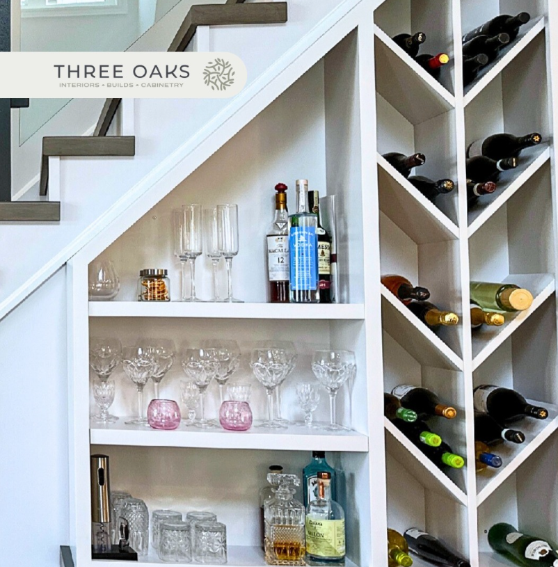 Built-in white shelving under a staircase, holding liquor bottles, glassware, and wine bottles arranged in a wine rack.