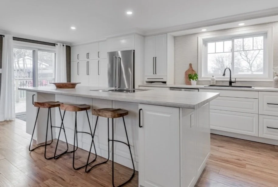 Bright modern kitchen with white cabinets, stainless steel refrigerator, and a large island with wooden stools. Large window over the sink provides natural light, wooden flooring throughout.