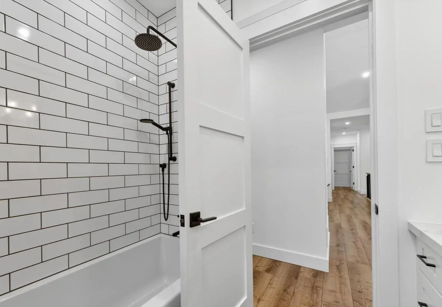 A bathroom with a bathtub and shower on the left, white subway tile on the wall, and a hallway with wooden flooring and white walls.
